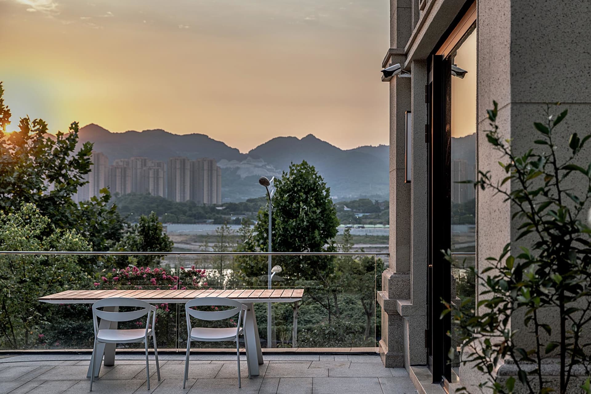 A minimalist balcony with wooden table, white chairs, glass railing, and concrete pillars overlooking mountains and cityscape