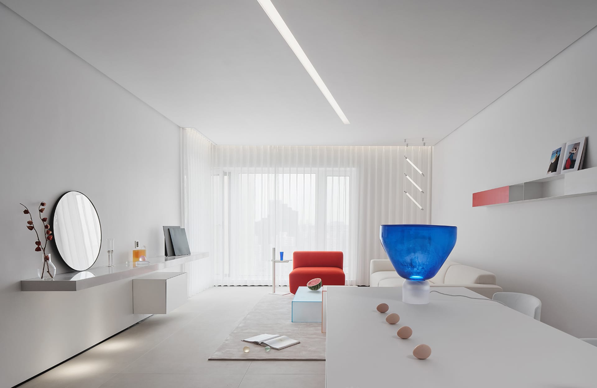 A minimalist living room featuring a glass table illuminated by blue lighting, with blinds allowing natural light in.