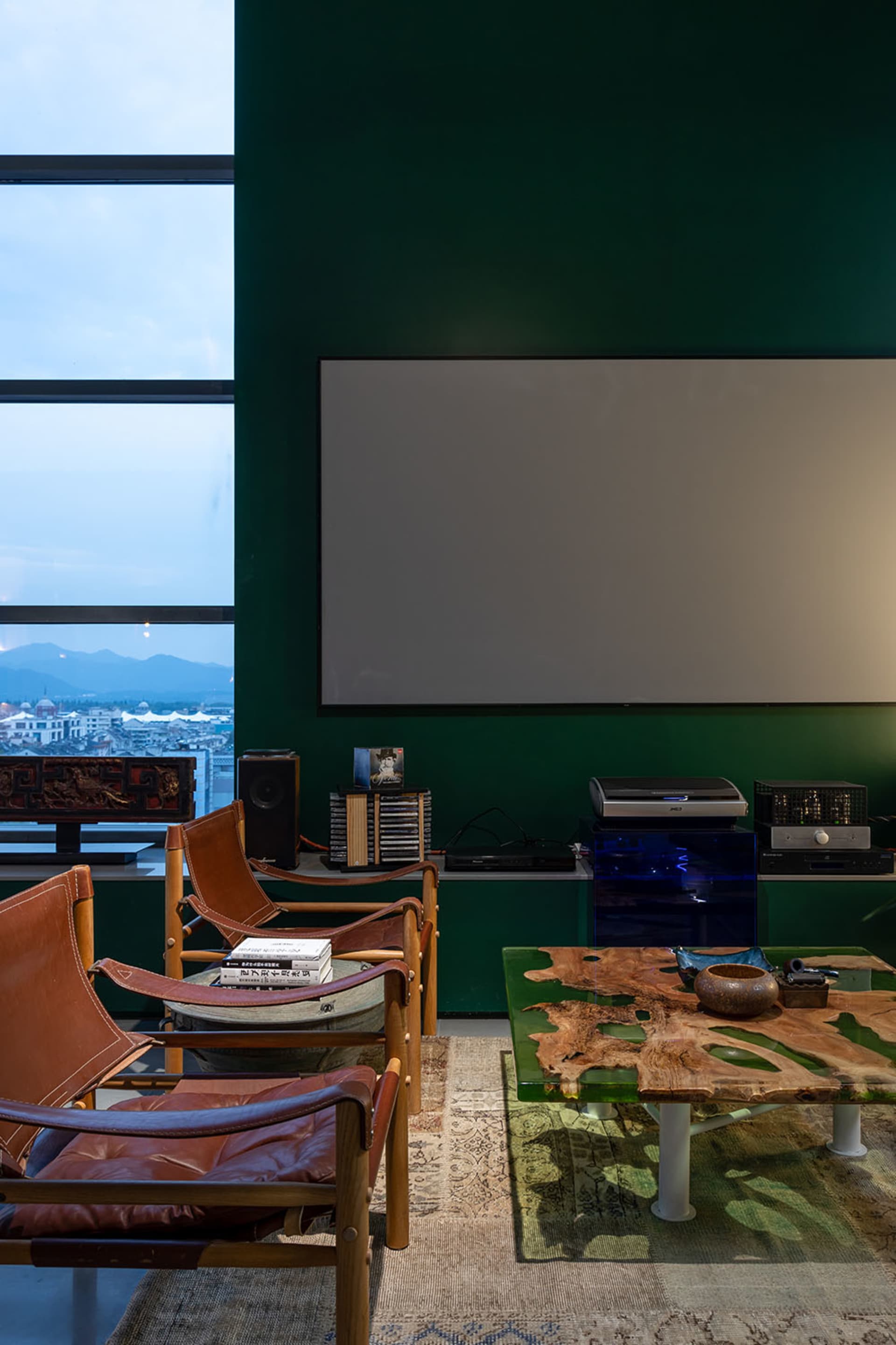 A modern living room with leather chairs, a coffee table, and a large screen on a green wall, featuring concrete flooring and