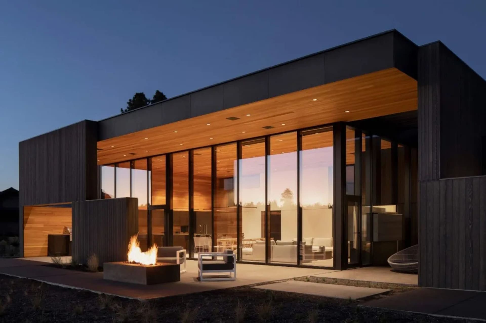 Modern residential exterior with wooden paneling and glass, featuring a fire pit and outdoor furniture at dusk.