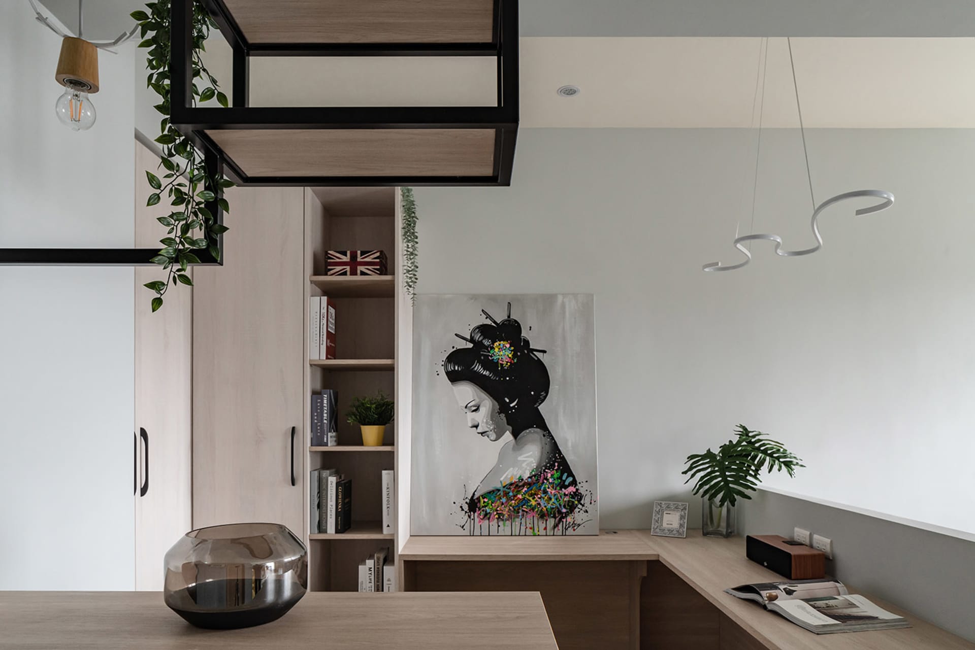A modern industrial office space featuring wooden furniture, a hanging plant, a geometric light fixture, and a painting of a