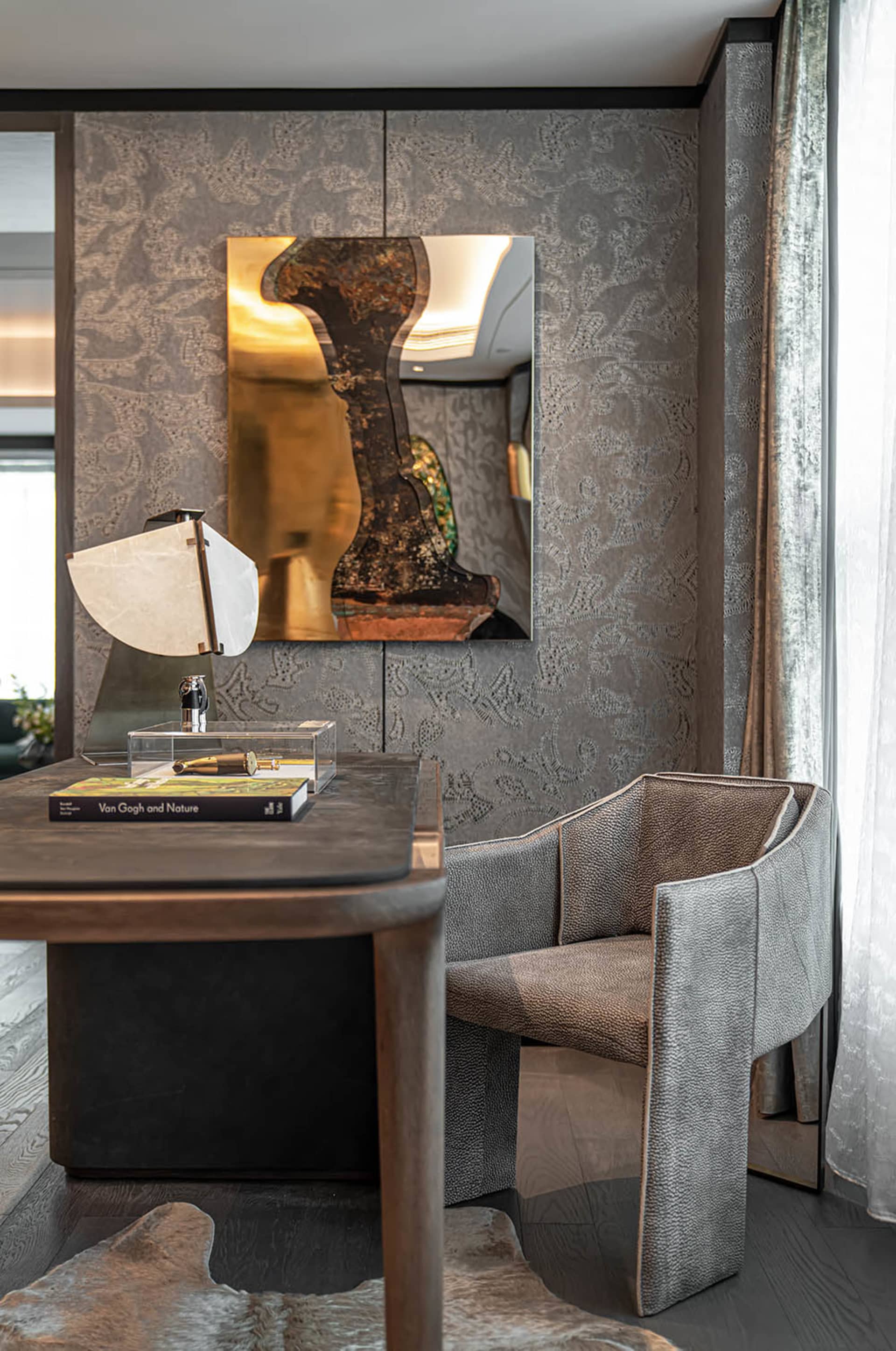A minimalist office featuring a wooden desk, a leather-covered armchair, and a fabric-covered curtain, with a lamp and a book