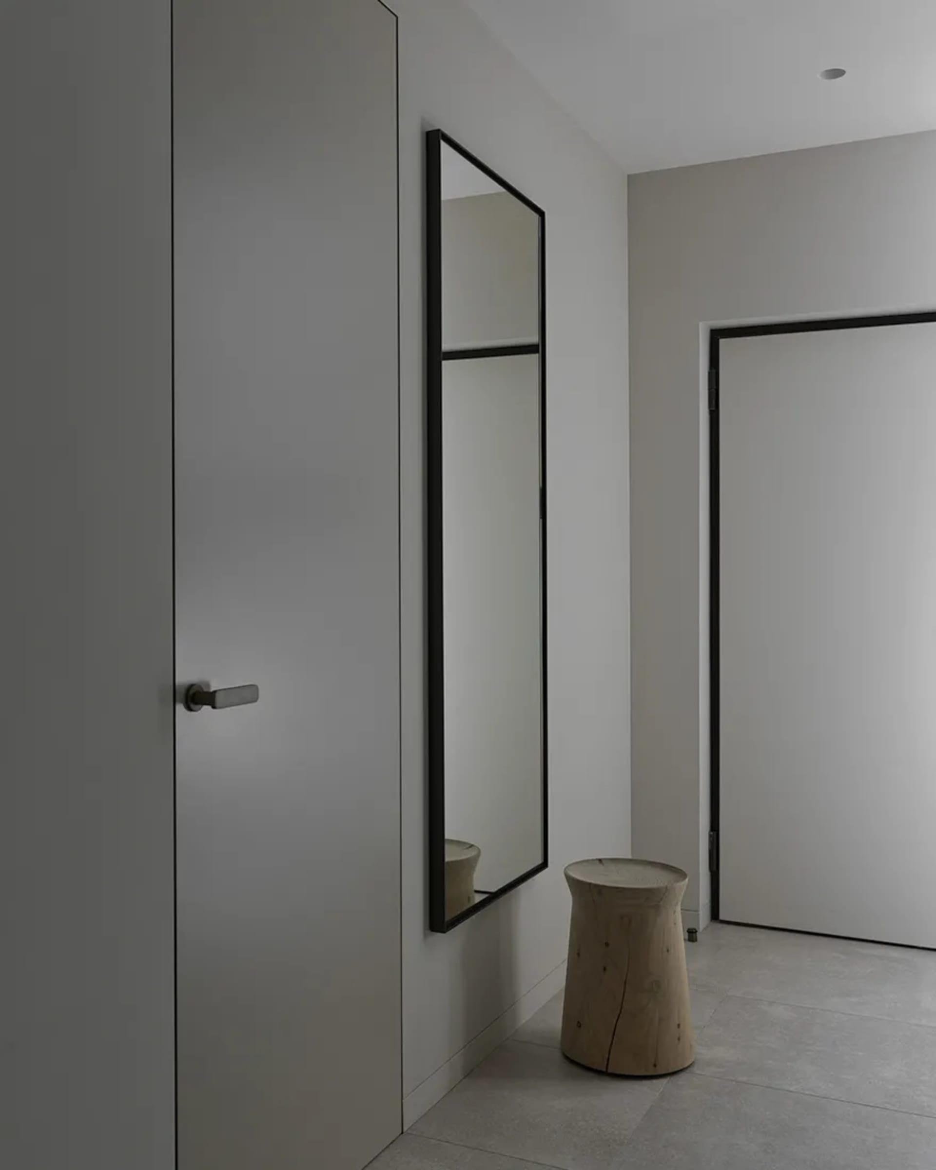A minimalist entryway featuring a wooden stool, white walls, and a large mirror with a black frame.
