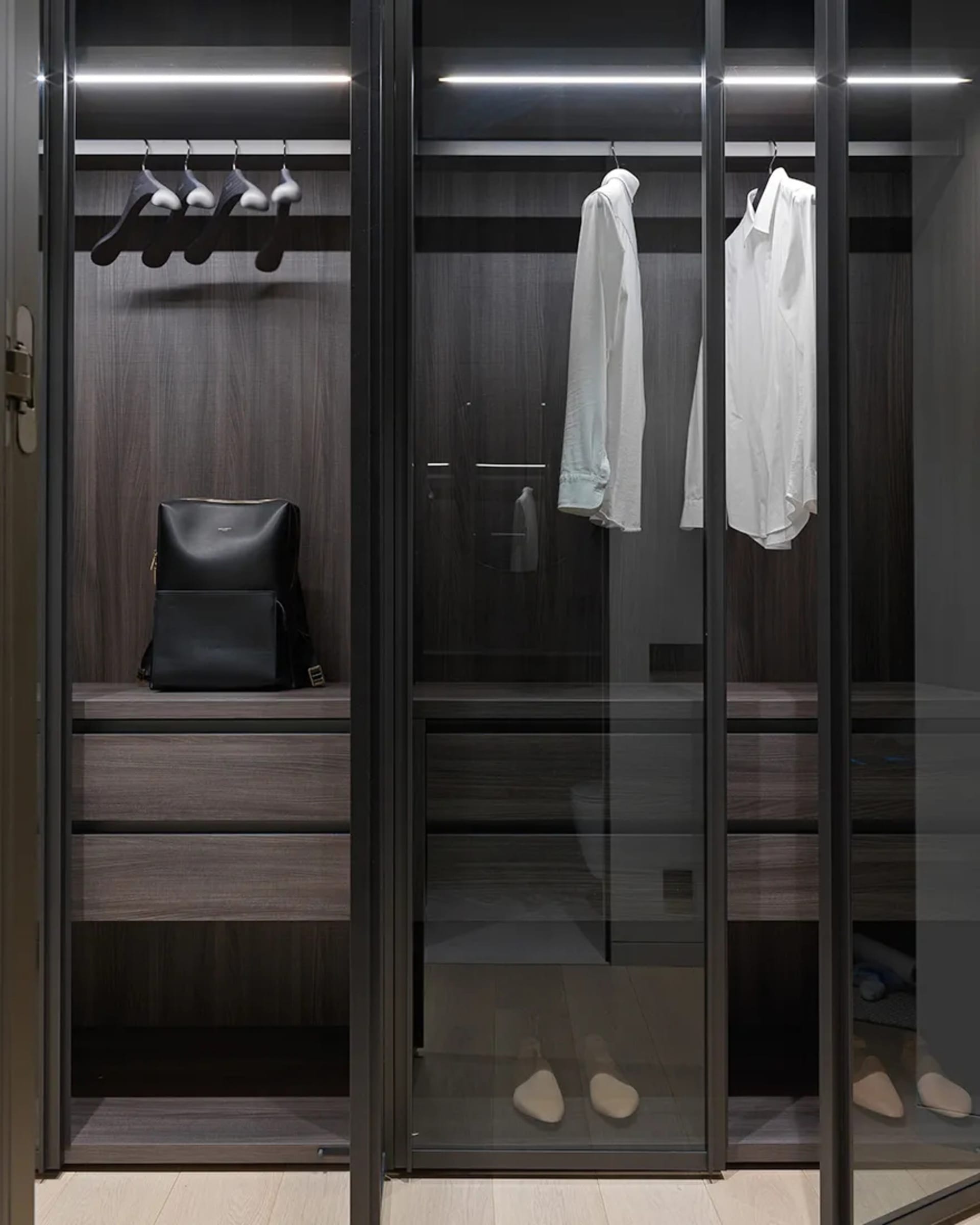 Modern dark wood metallic closet with glass doors, hanging clothes, and shoe rack.