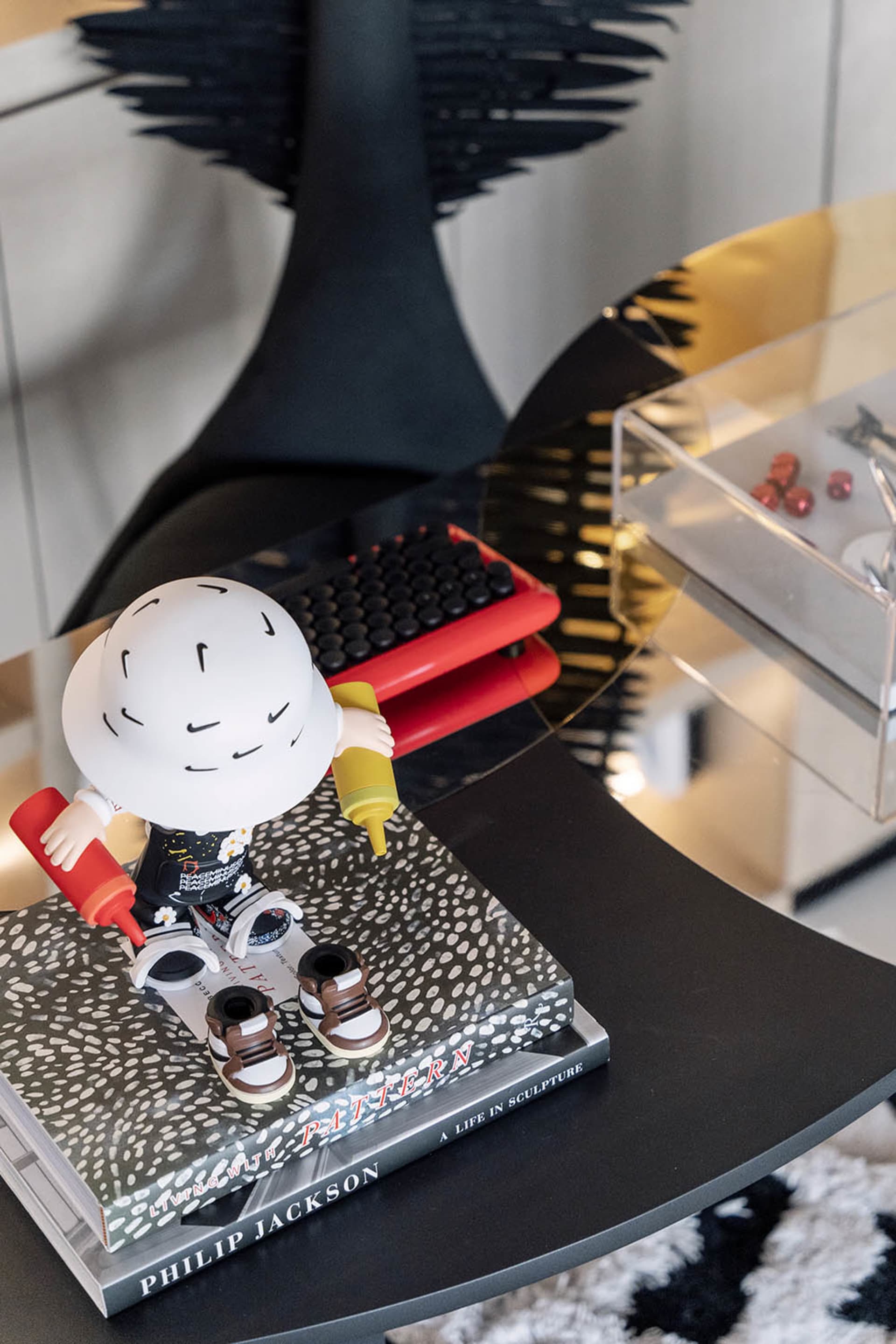 A modern interior design featuring a black round table with a patterned book, a toy figure holding ketchup bottle, and a clea