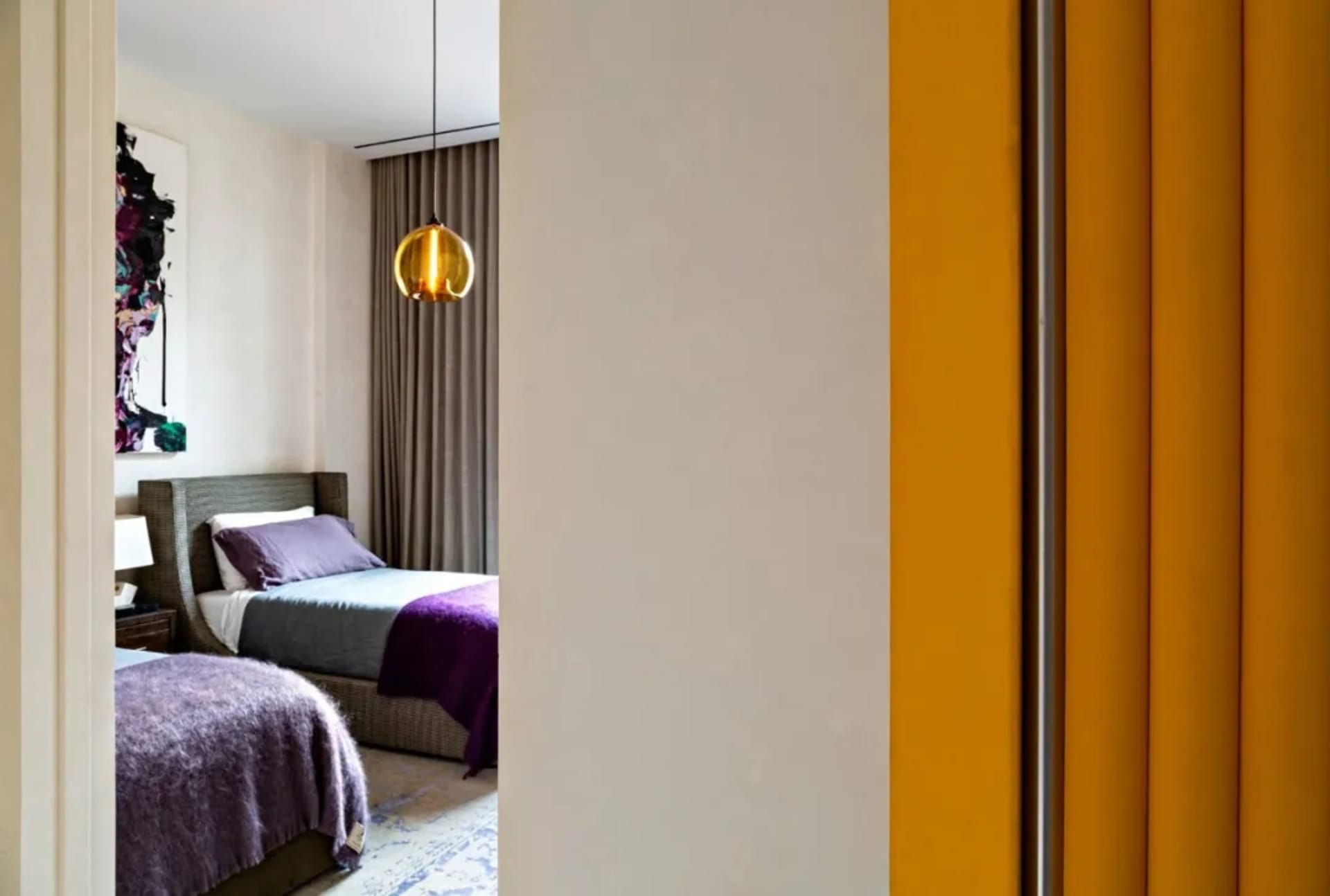A modern bedroom with a purple blanket on the bed, featuring a wicker bed frame, a glass pendant light, and grey and purple b