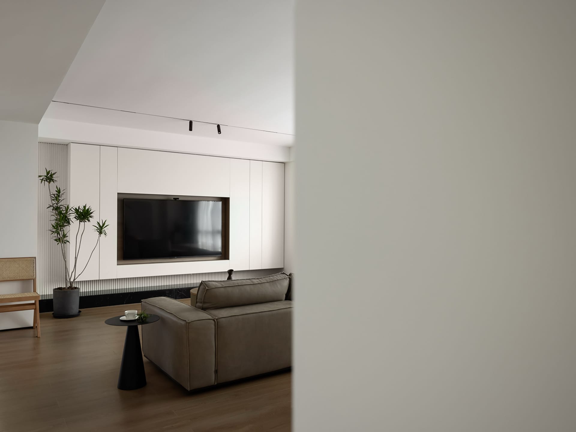 A modern minimalist living room featuring a grey sectional sofa, a black side table, a flat-screen TV embedded in a white pan
