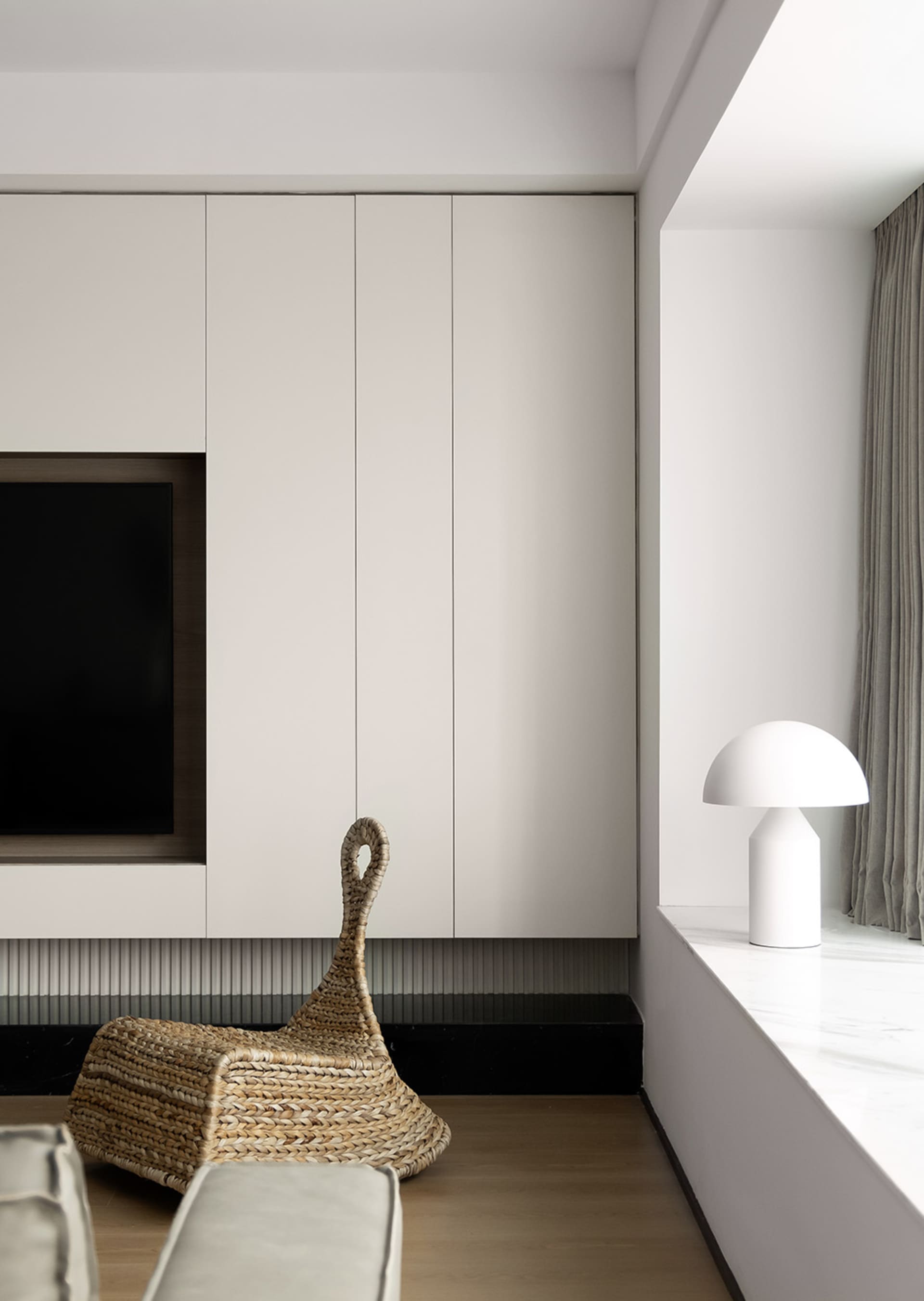 A modern minimalist living room with light-colored furniture, white cabinets, and a wicker bag on the floor.