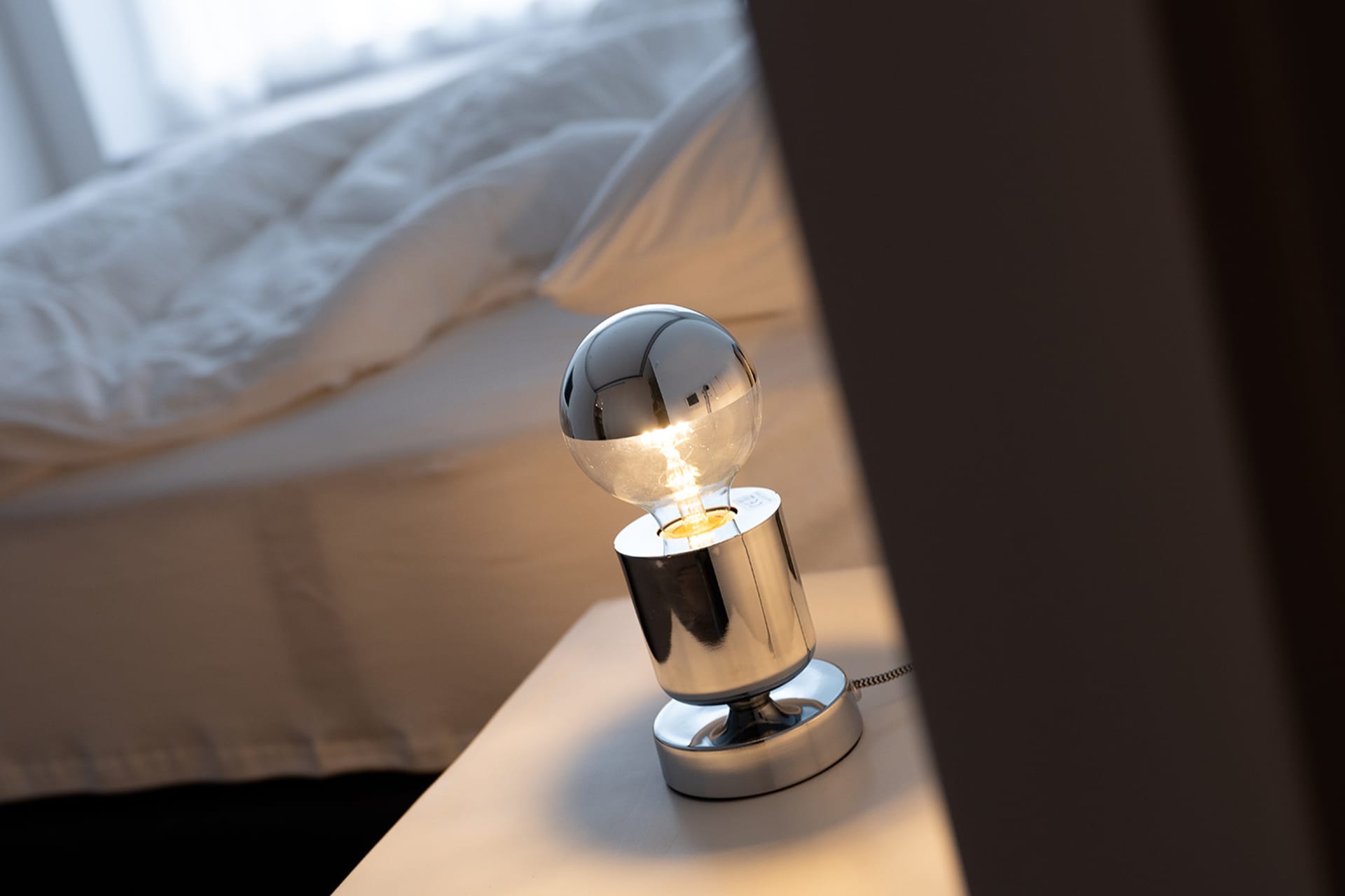 A minimalist bedroom featuring a metallic table lamp with a glowing lightbulb and a neatly made bed with white bedding.