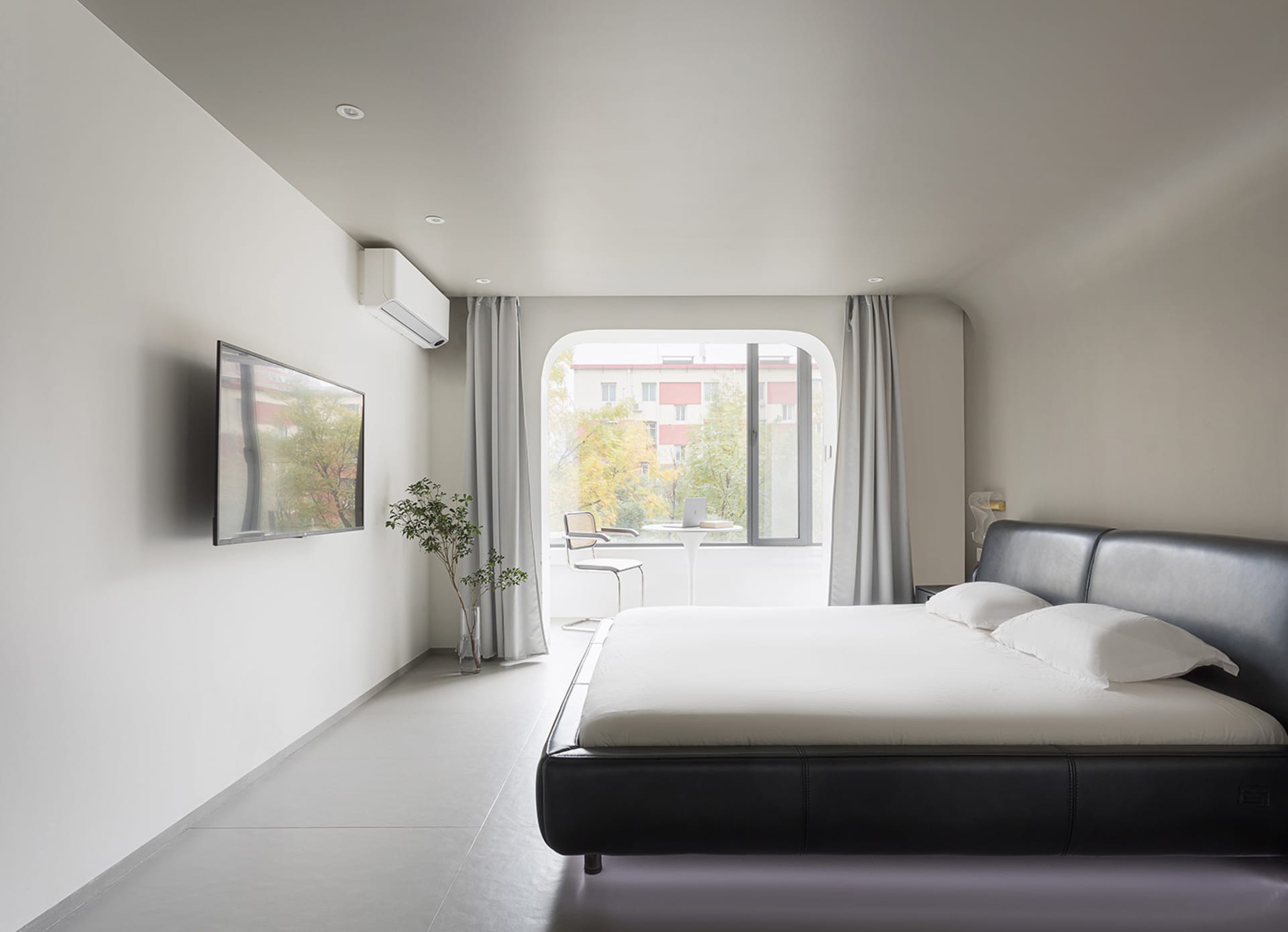 A modern bedroom featuring a white fabric bed, minimalistic decor, and a large window with a view of trees and buildings.