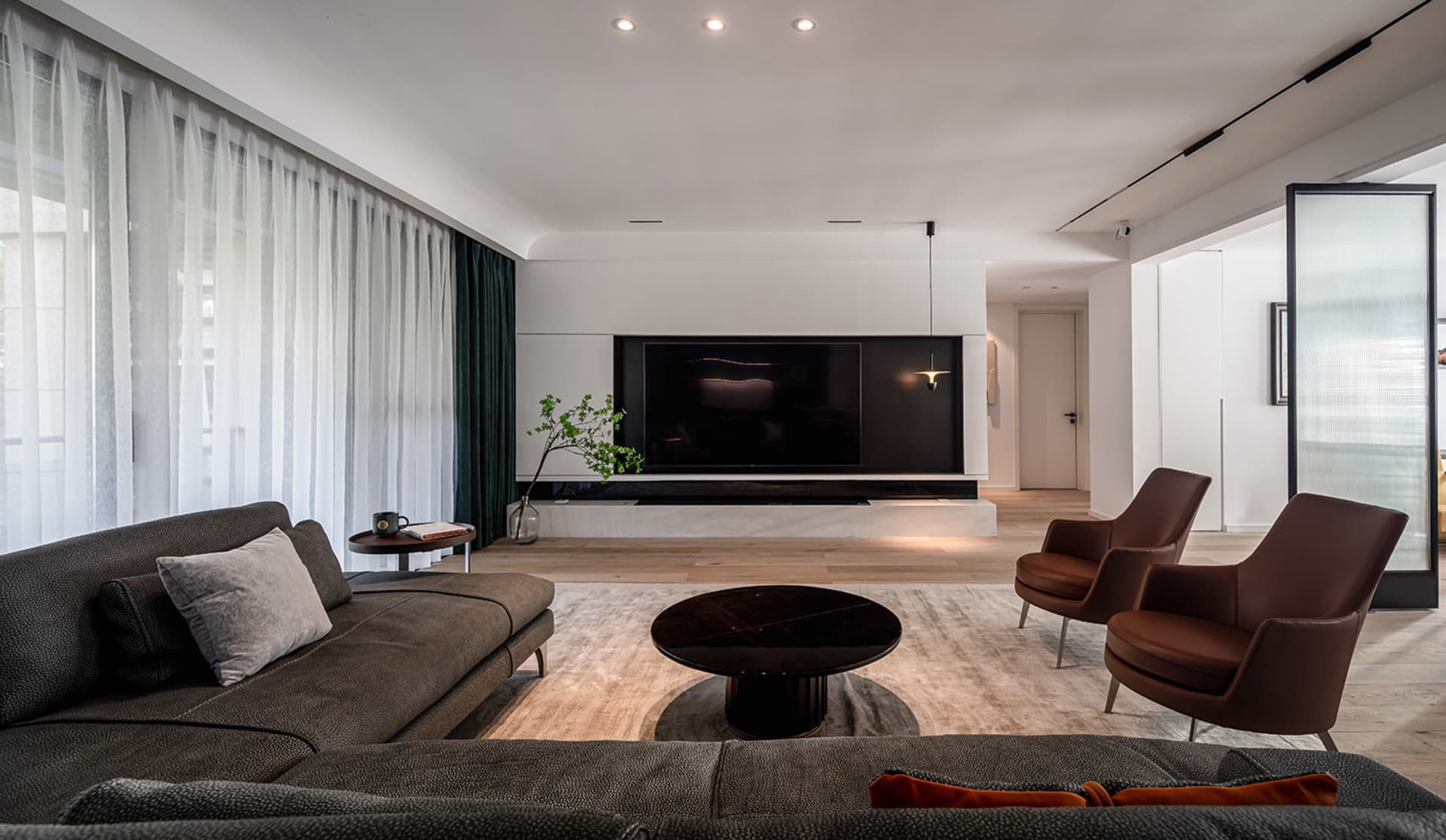 A modern living room with a gray sectional sofa, two brown armchairs, a round coffee table, and a large TV on the back wall,