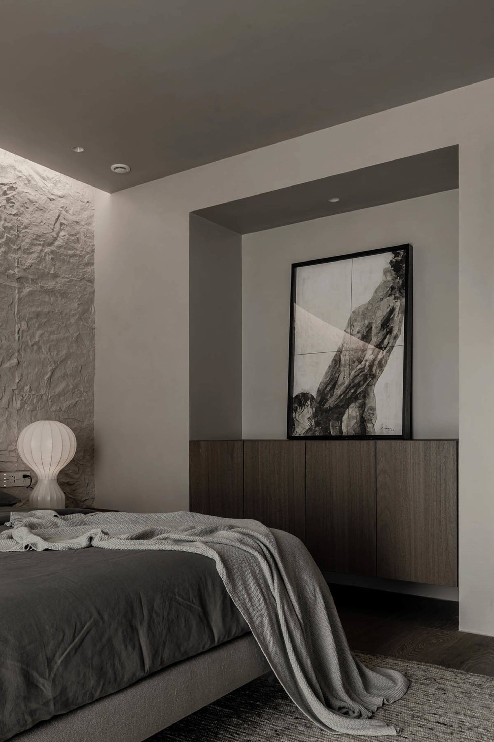 A minimalist bedroom with a textured stone wall, a bed with gray bedding, and a framed artwork.