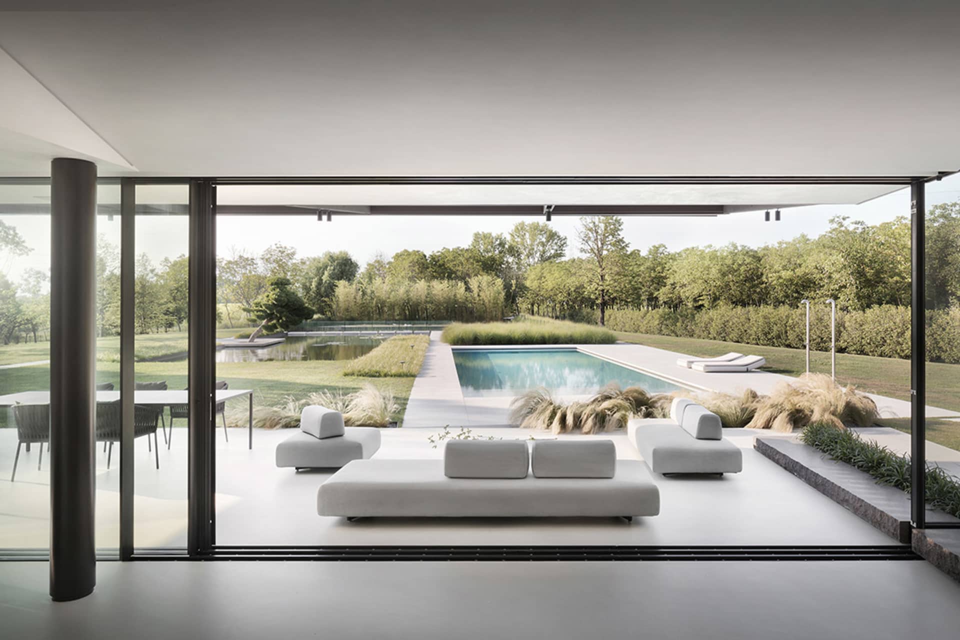 Modern residential living area with glass walls overlooking a pool surrounded by greenery.