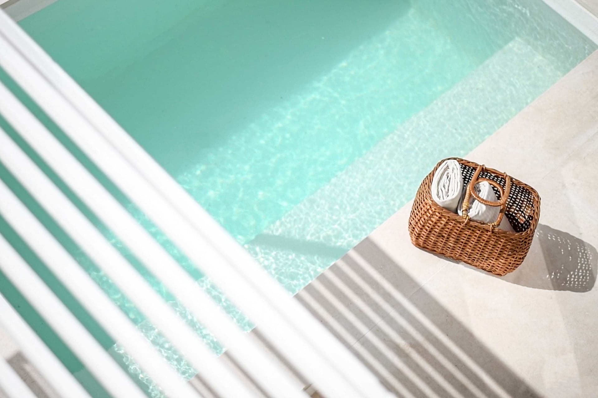 A wicker cane basket filled with rolled-up white towels placed beside a mint green swimming pool.
