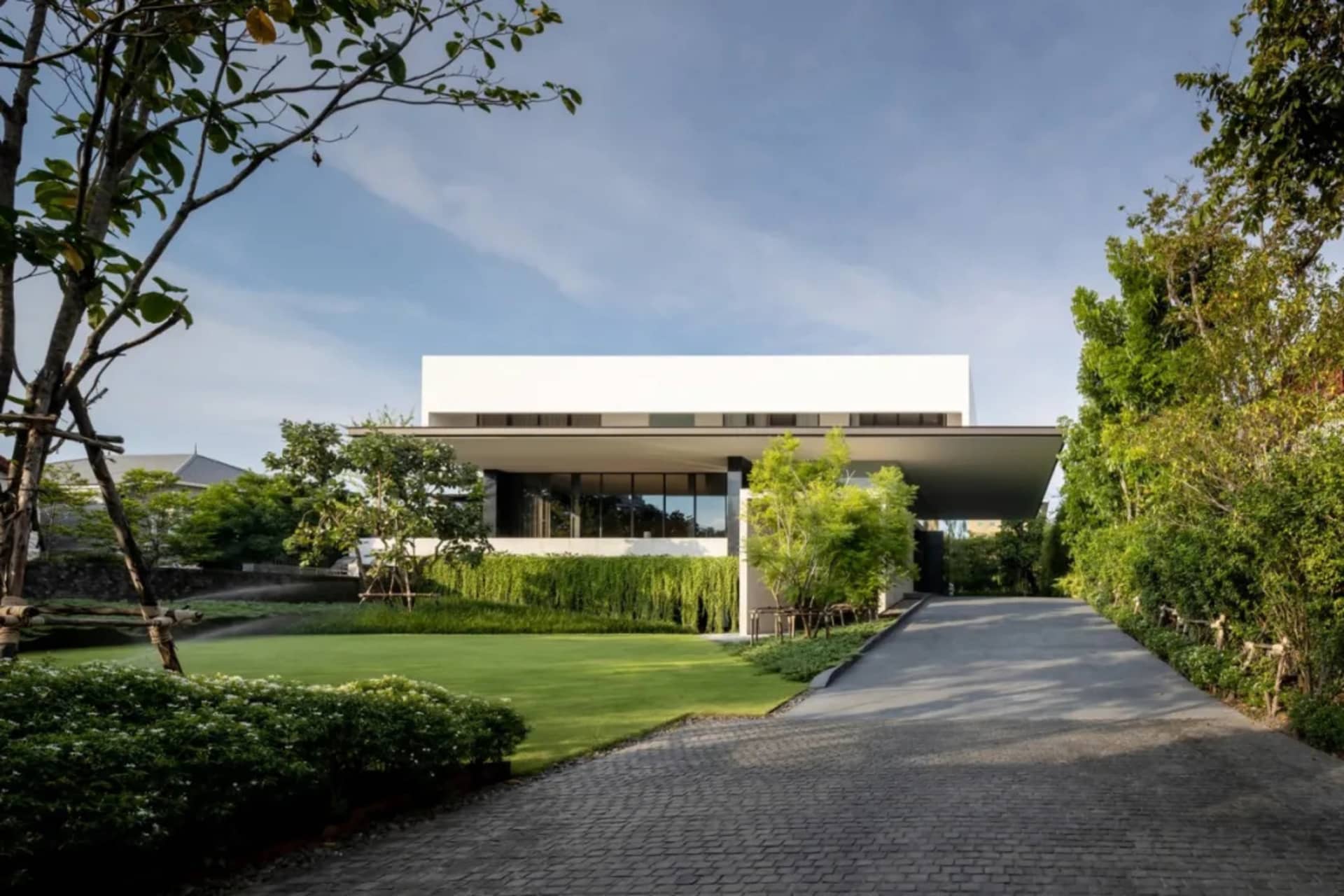 A modern residential home with a sleek, minimalist design featuring white concrete and glass materials, surrounded by lush gr