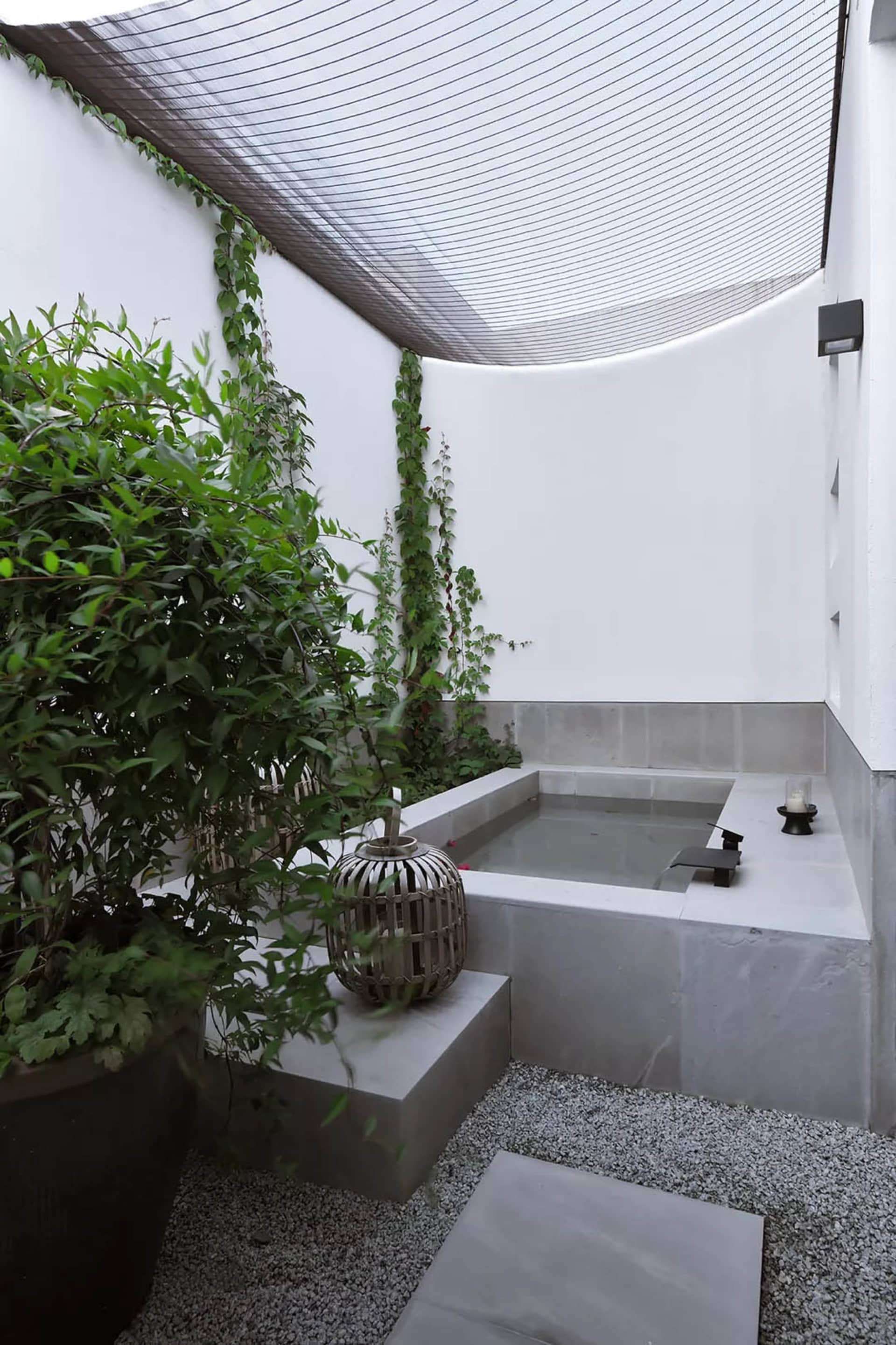 An outdoor Japanese-style bathtub made of concrete and stone, surrounded by greenery and a shaded canopy.
