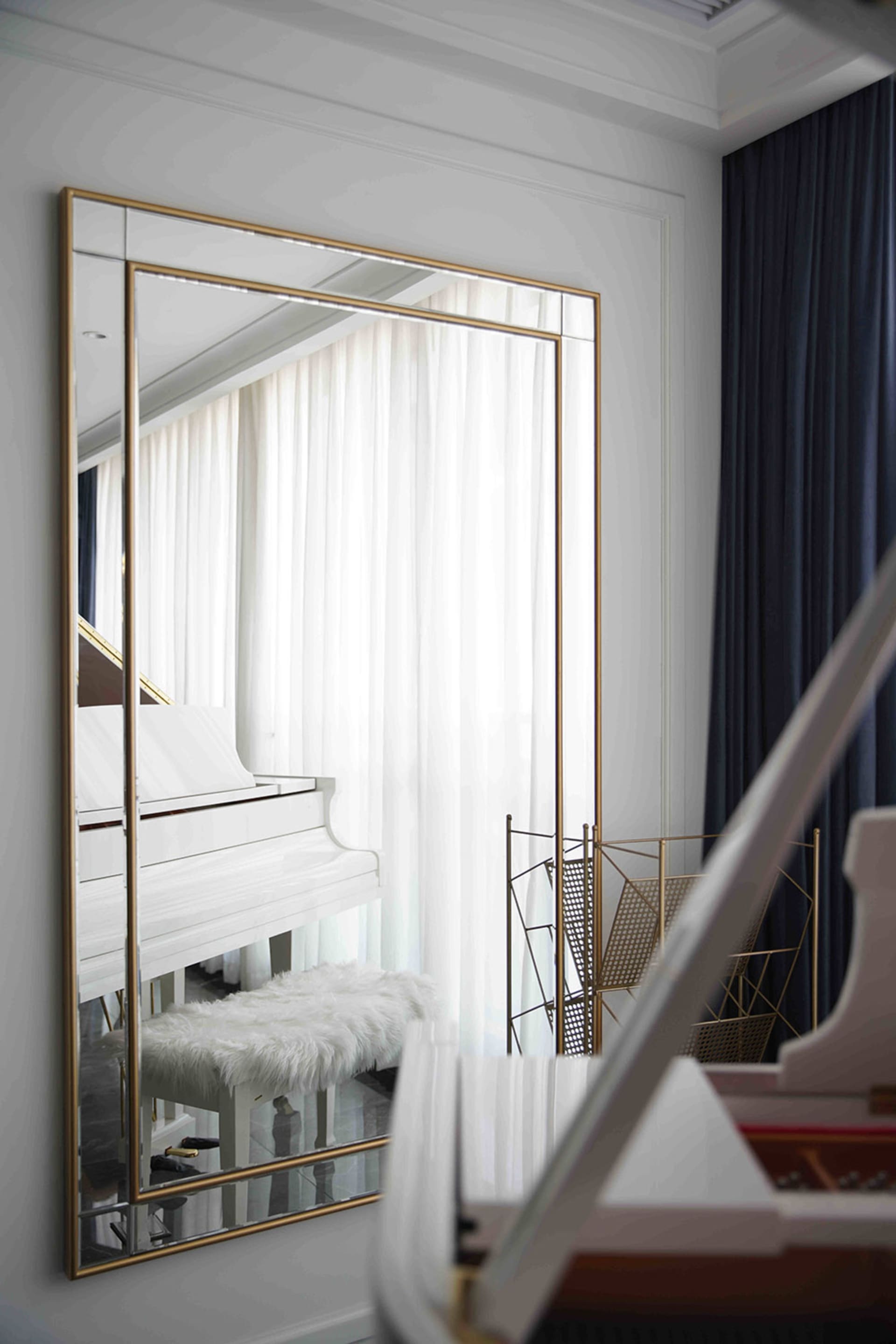 Glass-frame mirror reflecting a piano and a white curtain with a furry white cushion in a music room setting.