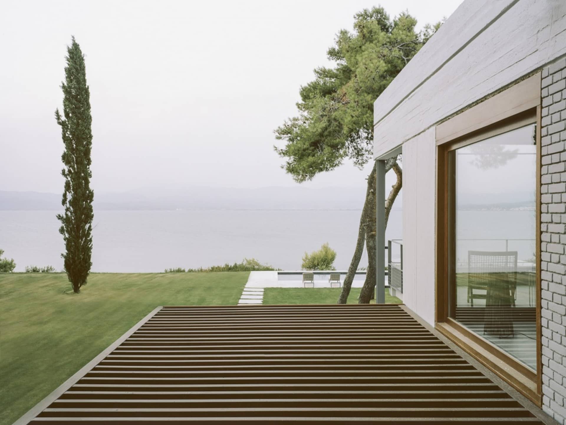 Modern house with wooden deck, glass windows, and concrete patio overlooking a calm lake.