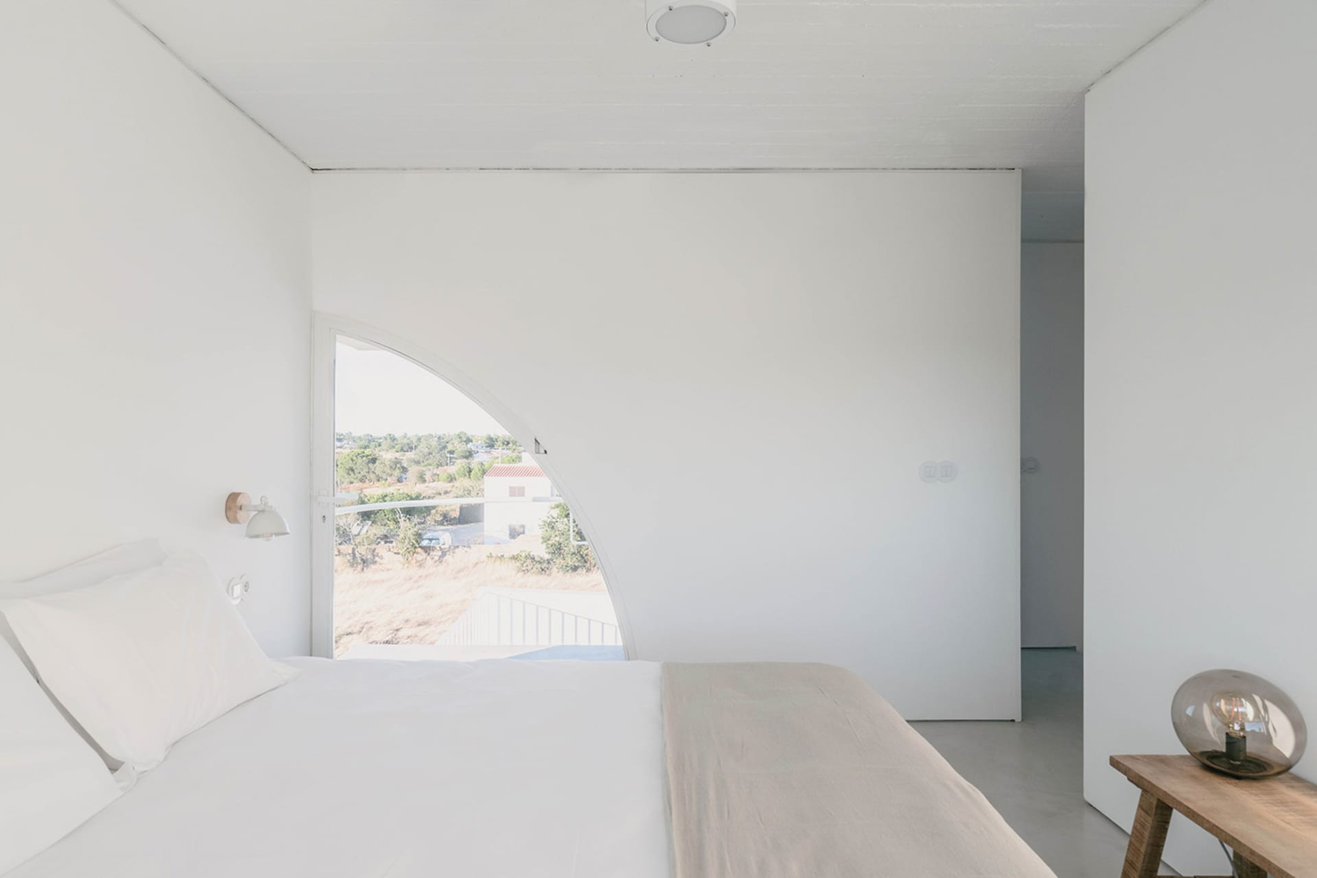 Modern minimalist bedroom with concrete walls and floor, featuring a large curved window overlooking a natural landscape.
