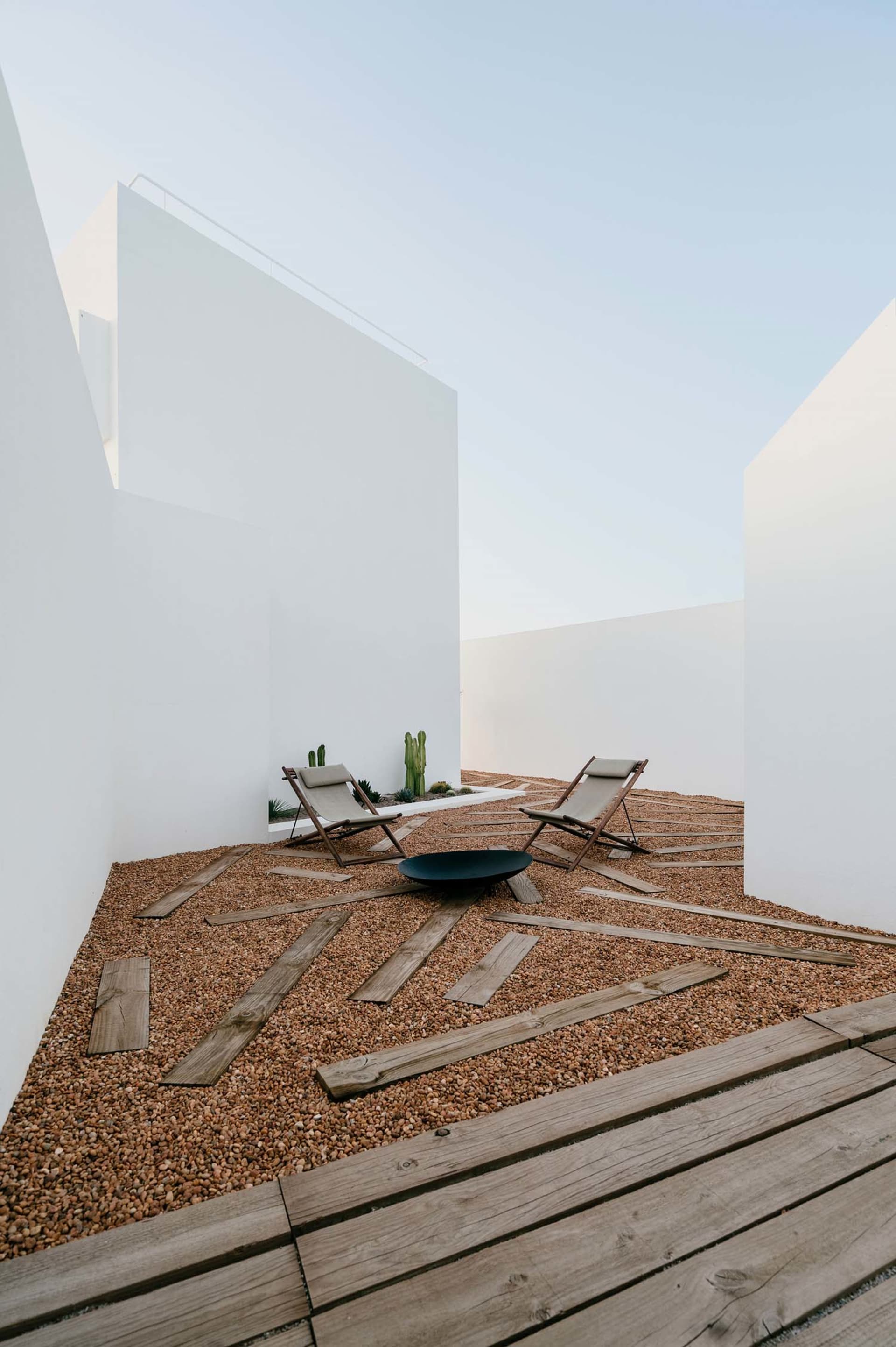 Two lounge chairs on a gravel floor surrounded by wooden planks, concrete walls, and cactus plants.