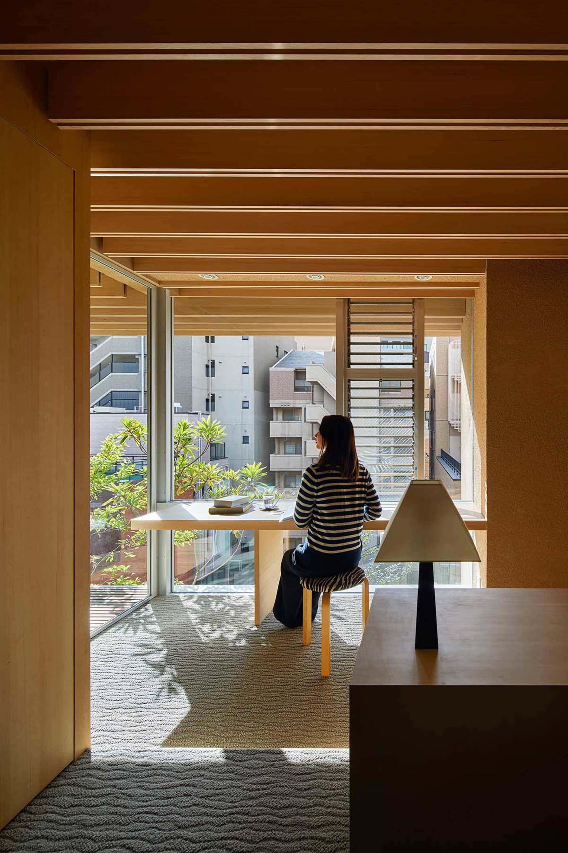 A person sitting at a wooden desk in a home office, with a chair and a lamp on a wooden table.