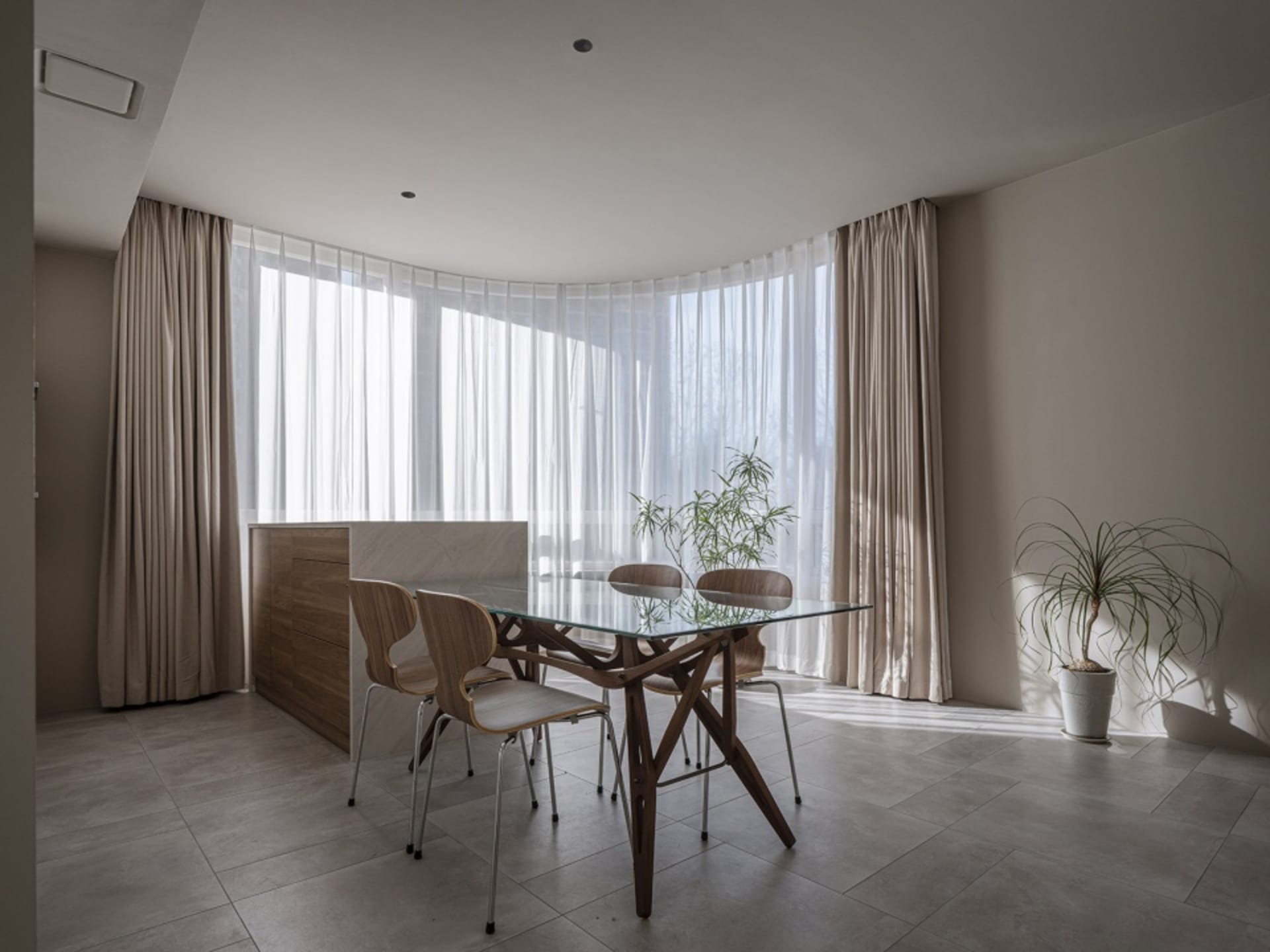 A modern dining room featuring a glass-top table, wooden chairs, and transparent curtains allowing natural light to flood the