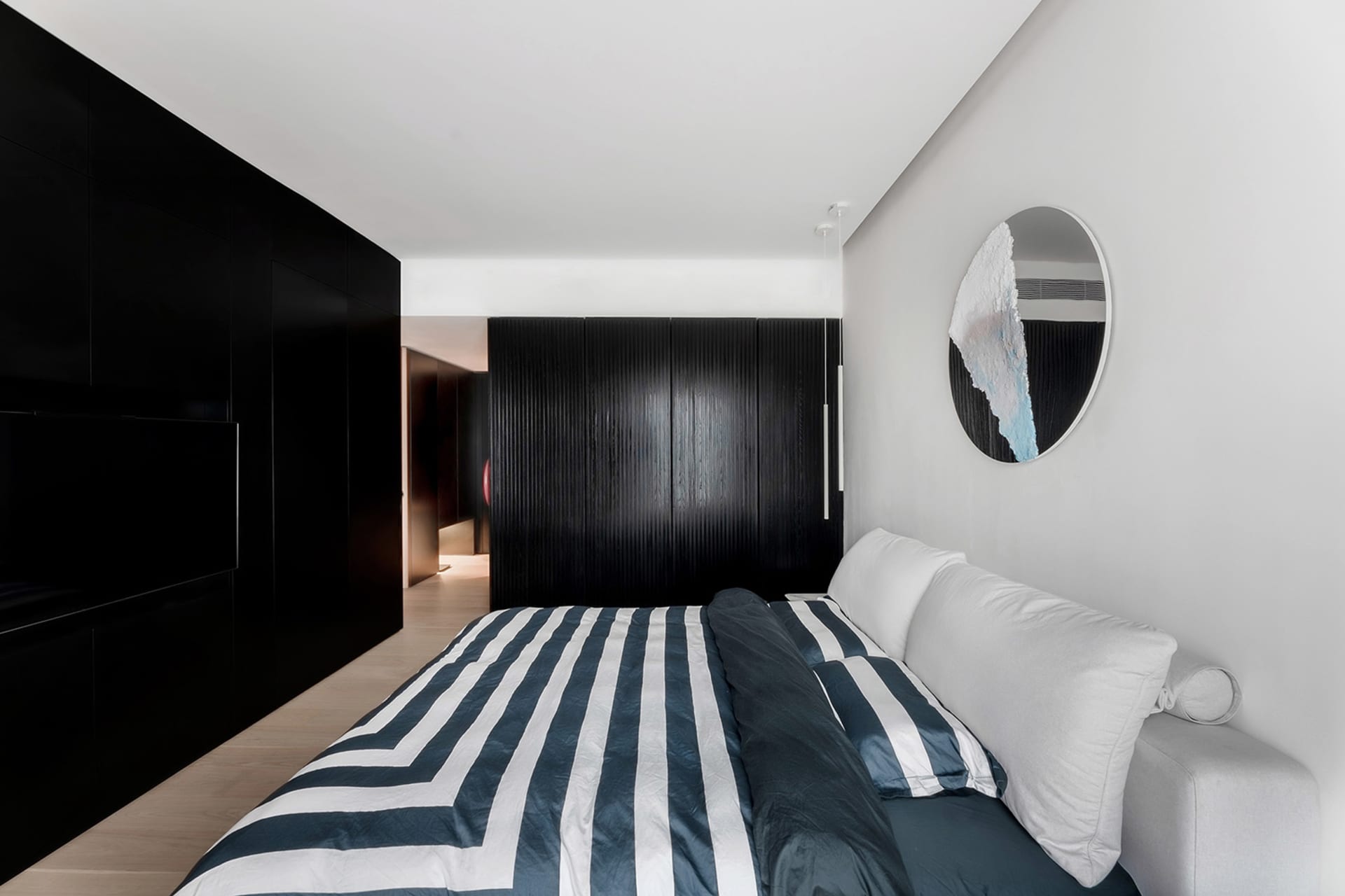 A modern minimalist bedroom with a striped blue and white bedspread, black cabinets, and a round mirror on the wall.