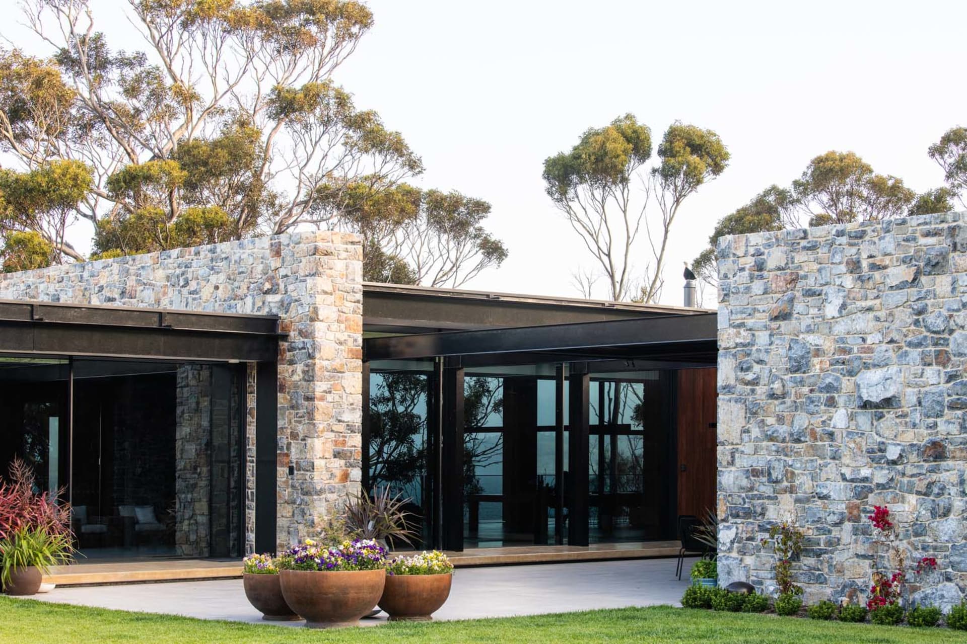 Modern residential exterior featuring stone and concrete walls, glass windows, and lush green landscaping.