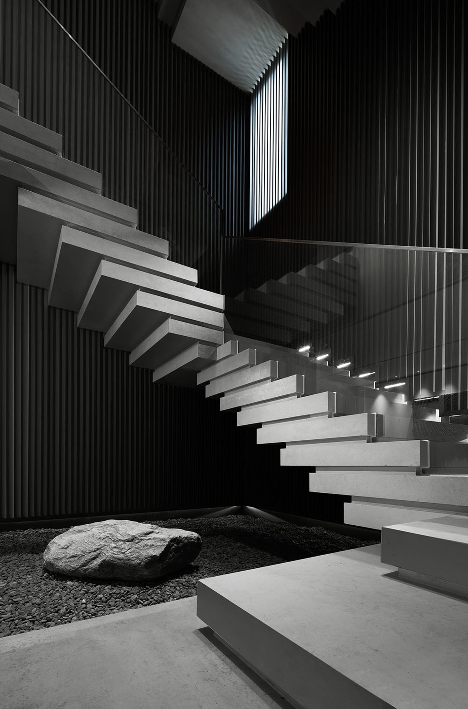 A modern staircase made of graphite concrete with a glass railing and a large rock in the foreground.