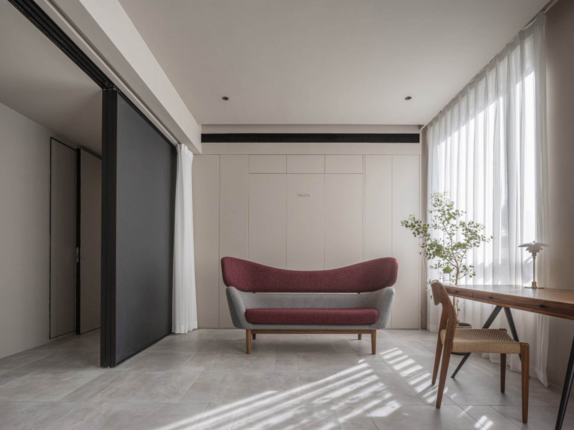 A minimalist living room with a modern sofa, wooden desk, and a plant by the window. The room features neutral-toned walls an