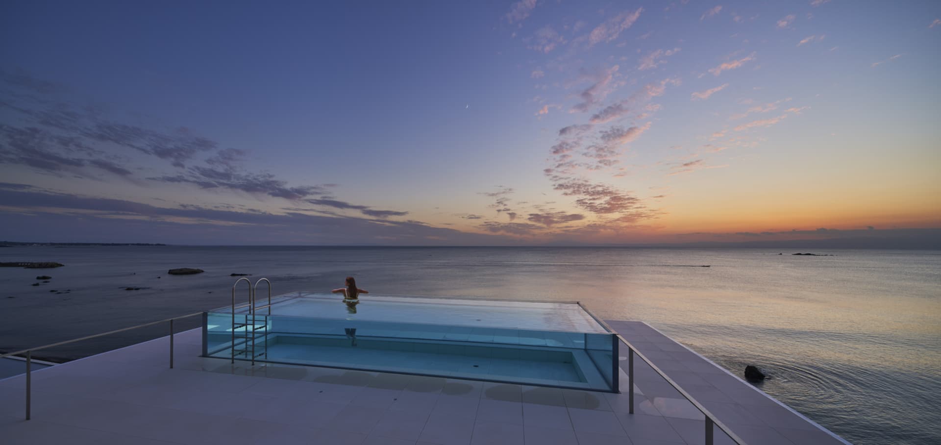A modern poolside view with a glass infinity pool overlooking the ocean during sunset.