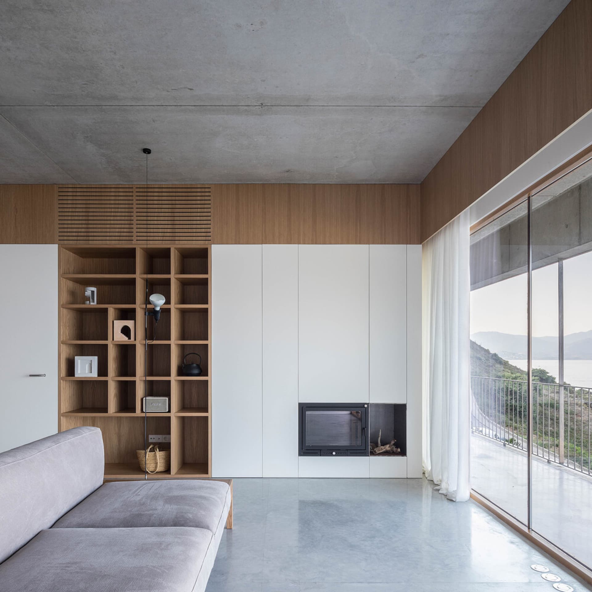 Modern minimalist residential interior featuring concrete, wood, and glass materials, with a white sofa, wooden shelves, and