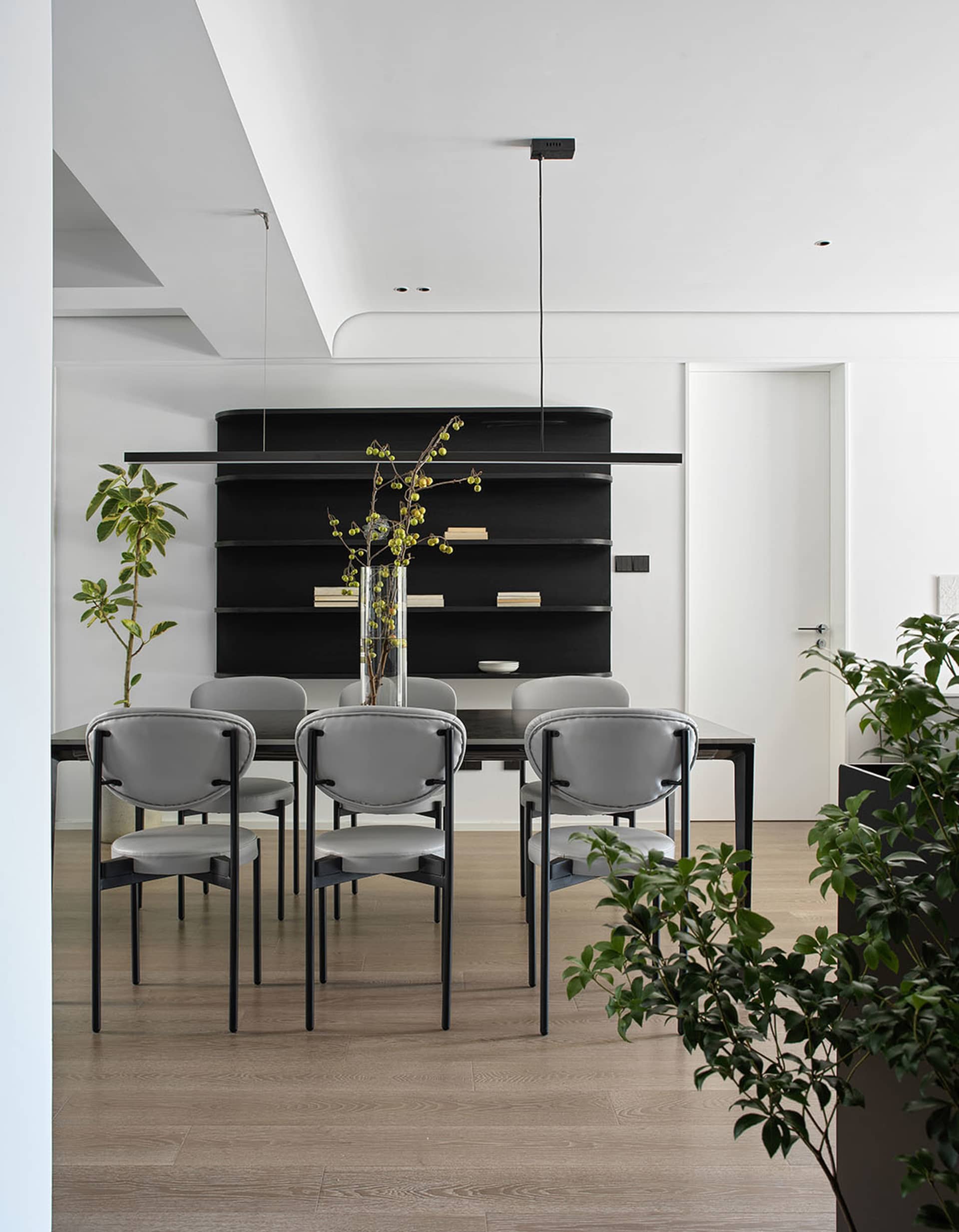 A modern dining room with a minimalist design, featuring a black table, white chairs, and a black shelving unit with books.