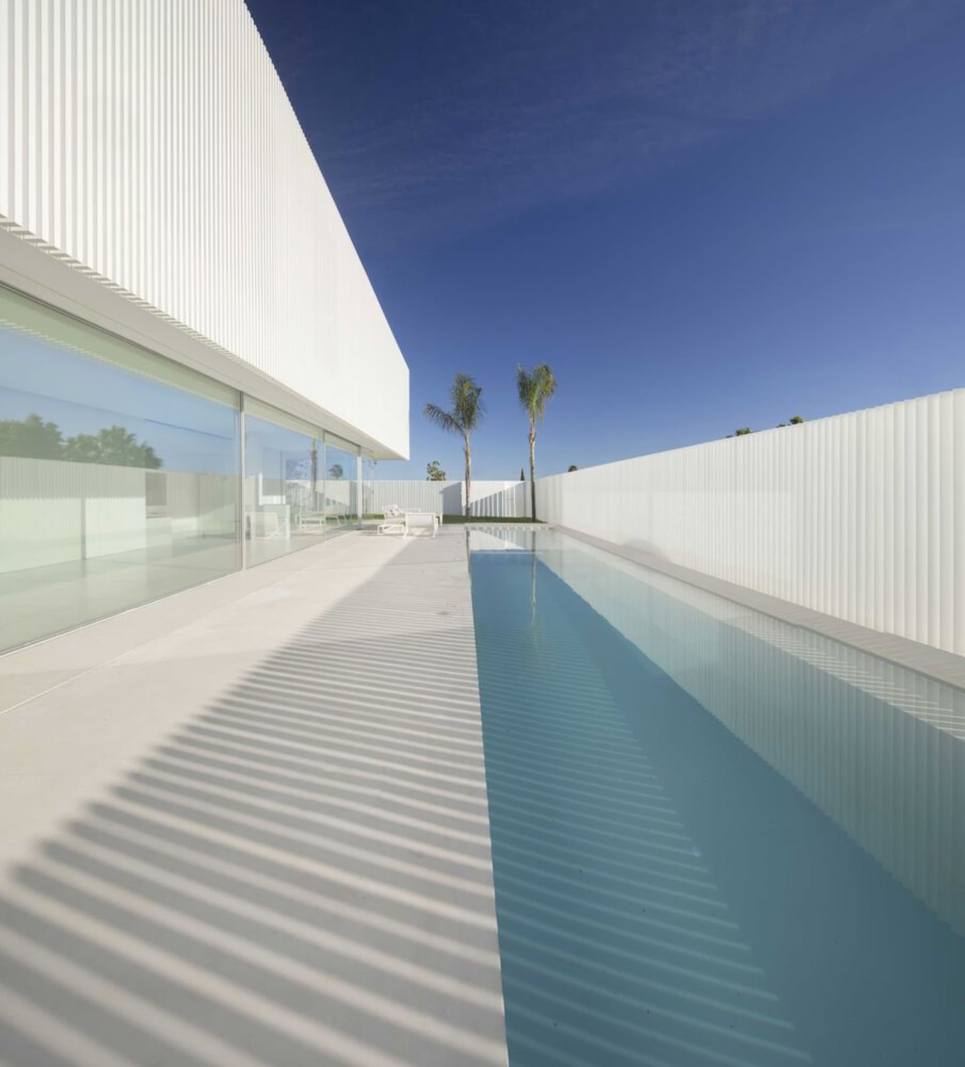 Modern white-rendered exterior of a private residence with a long pool and palm trees in the background.