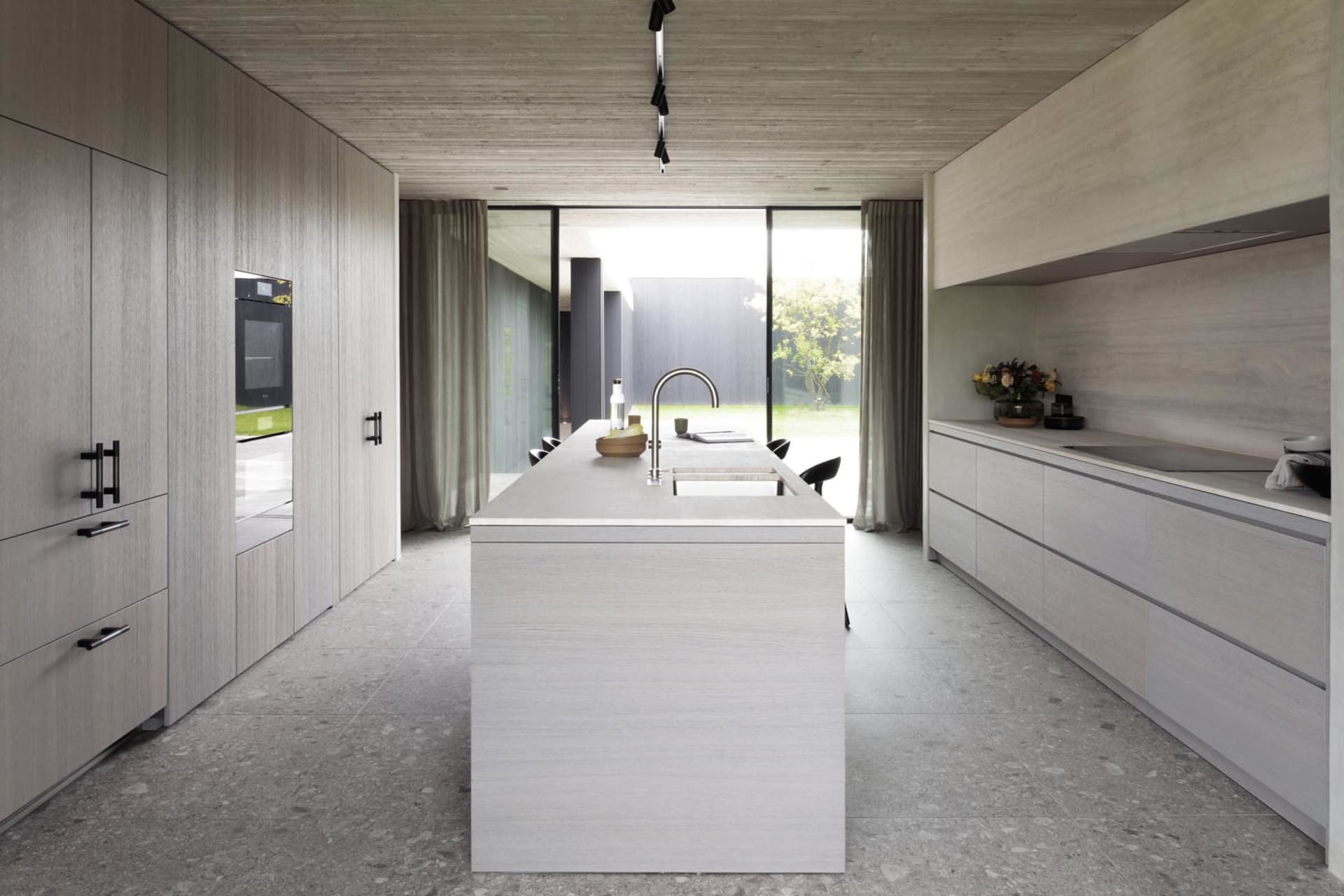 Modern, minimalist kitchen with concrete countertops and floor, featuring an island with a sink and a view of the outdoors th