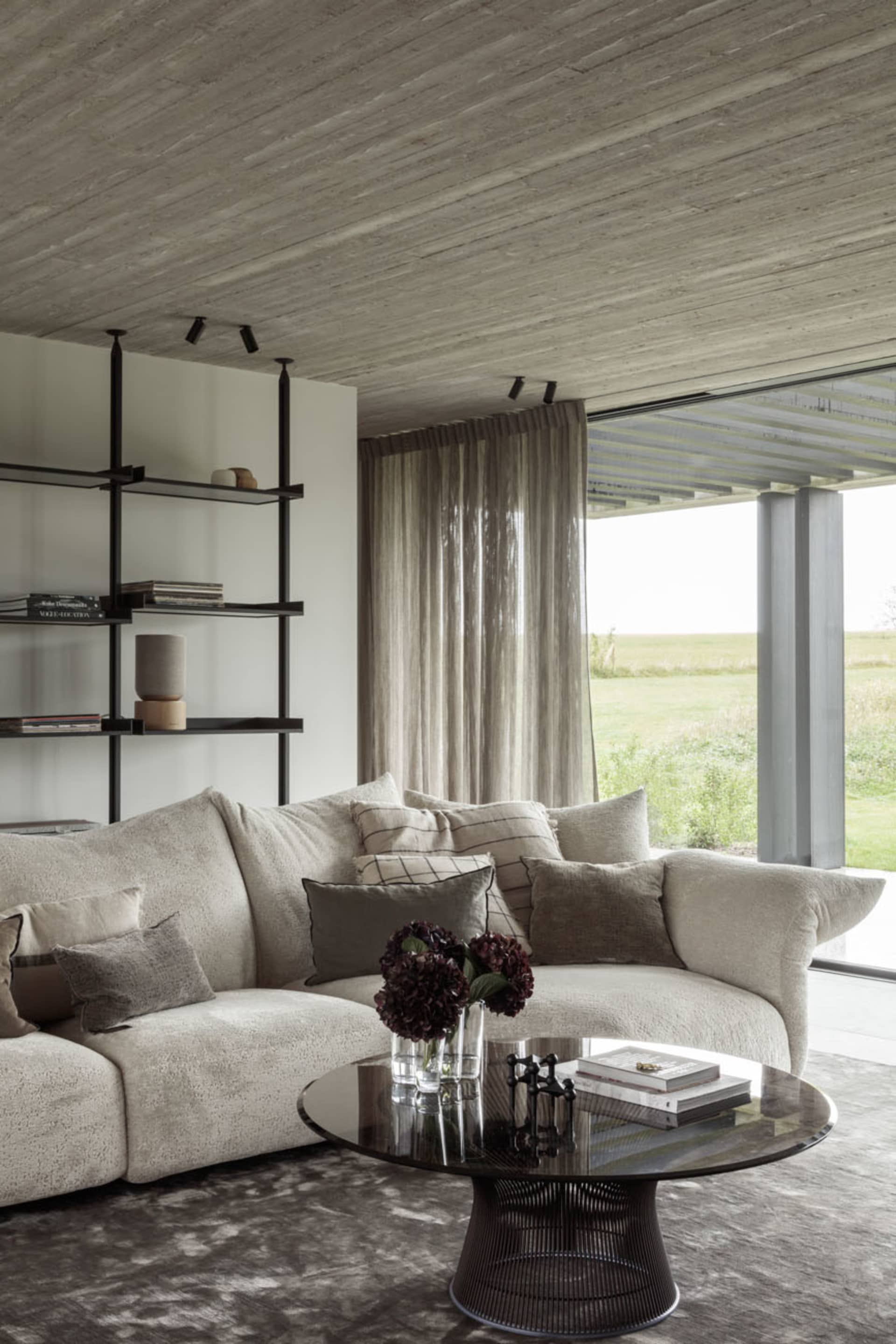 Modern living room with concrete ceiling, fabric sectional sofa, glass coffee table, and floor-to-ceiling windows showing gre