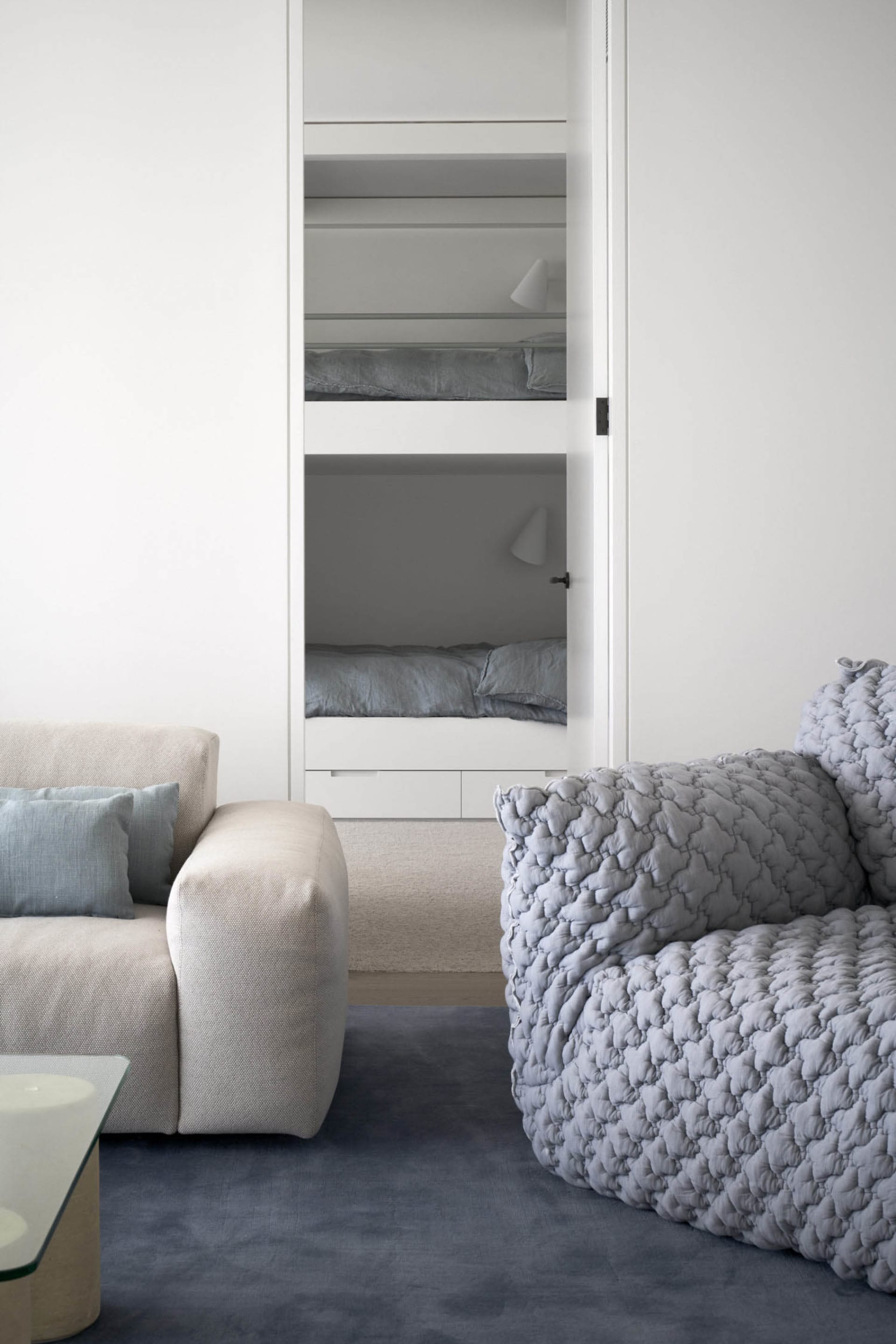 A minimalist living room with a beige sofa, a gray textured armchair, and a glass top table.