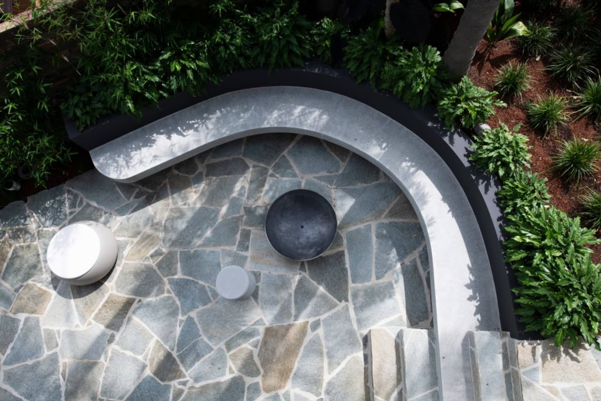 A modern patio featuring a curved granite bench and circular tables surrounded by green plants.