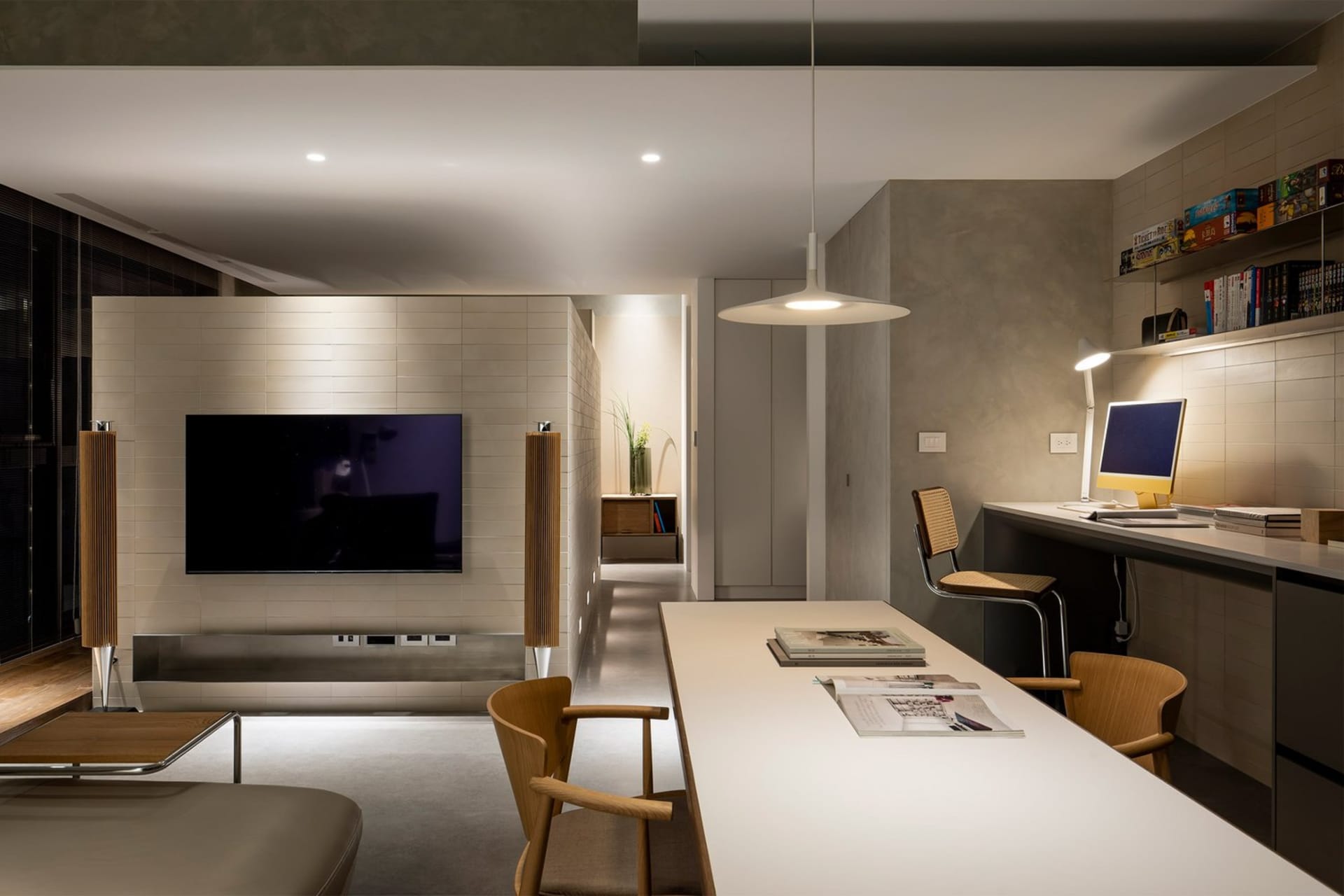 A minimalist living room with a concrete surface, featuring a wall-mounted TV, a long table with a computer setup, and a comf