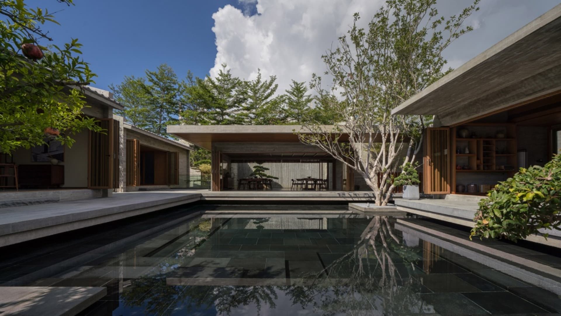 Modern residential pool with concrete walls and floors, surrounded by greenery, under a blue sky with clouds.