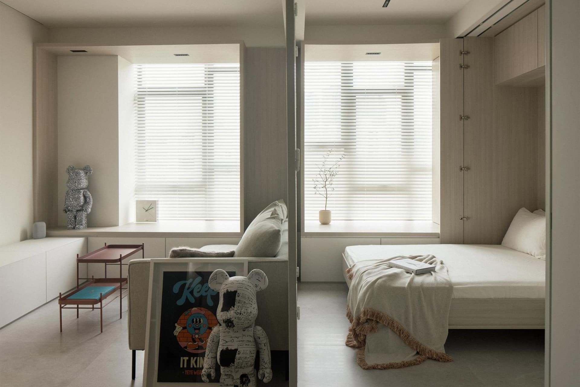 A minimalist bedroom with a bed, couch, and window with blinds. Room features a neutral color palette and simple furnishings.