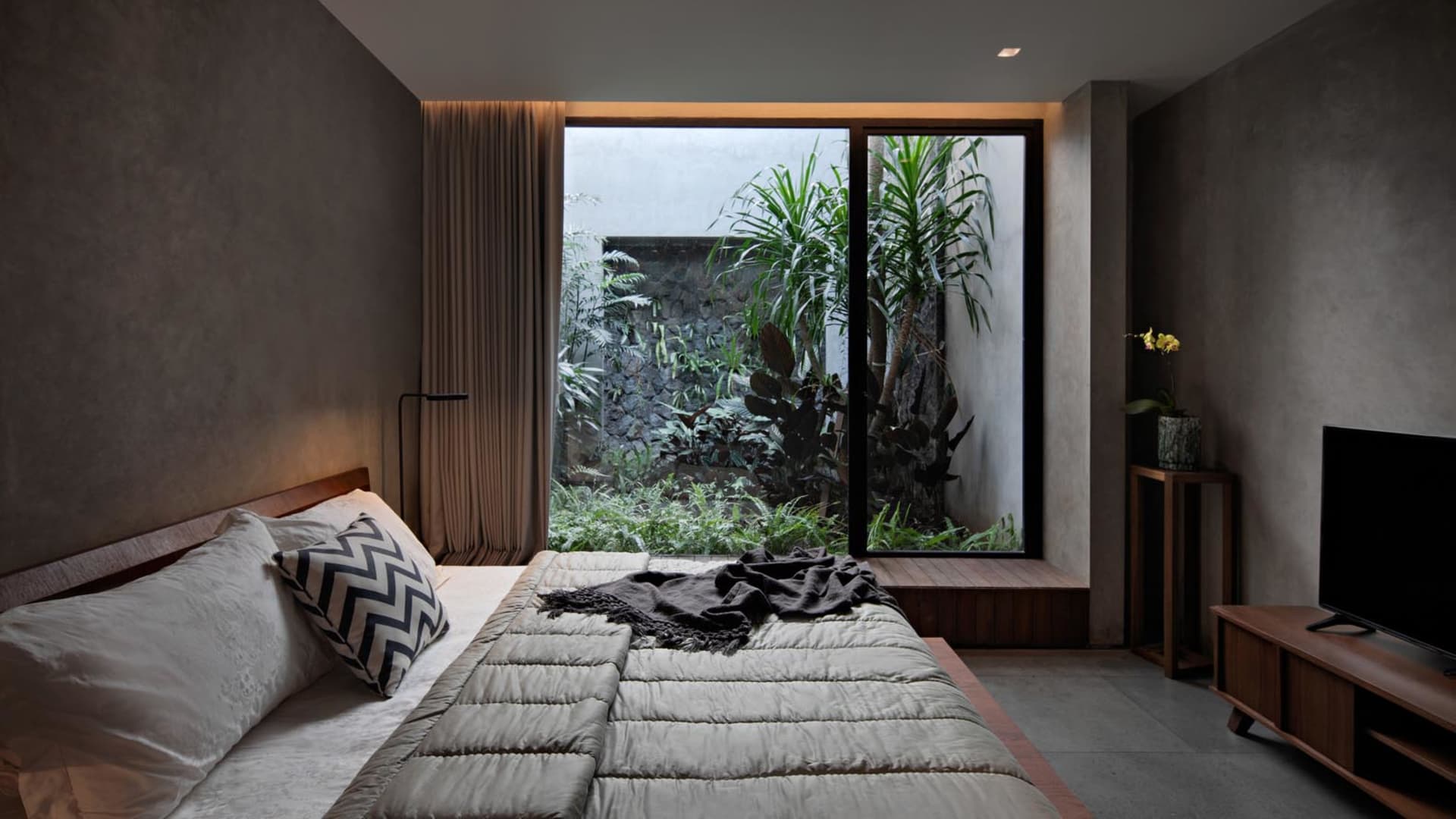 Modern bedroom with concrete walls, a large bed, and a garden view.
