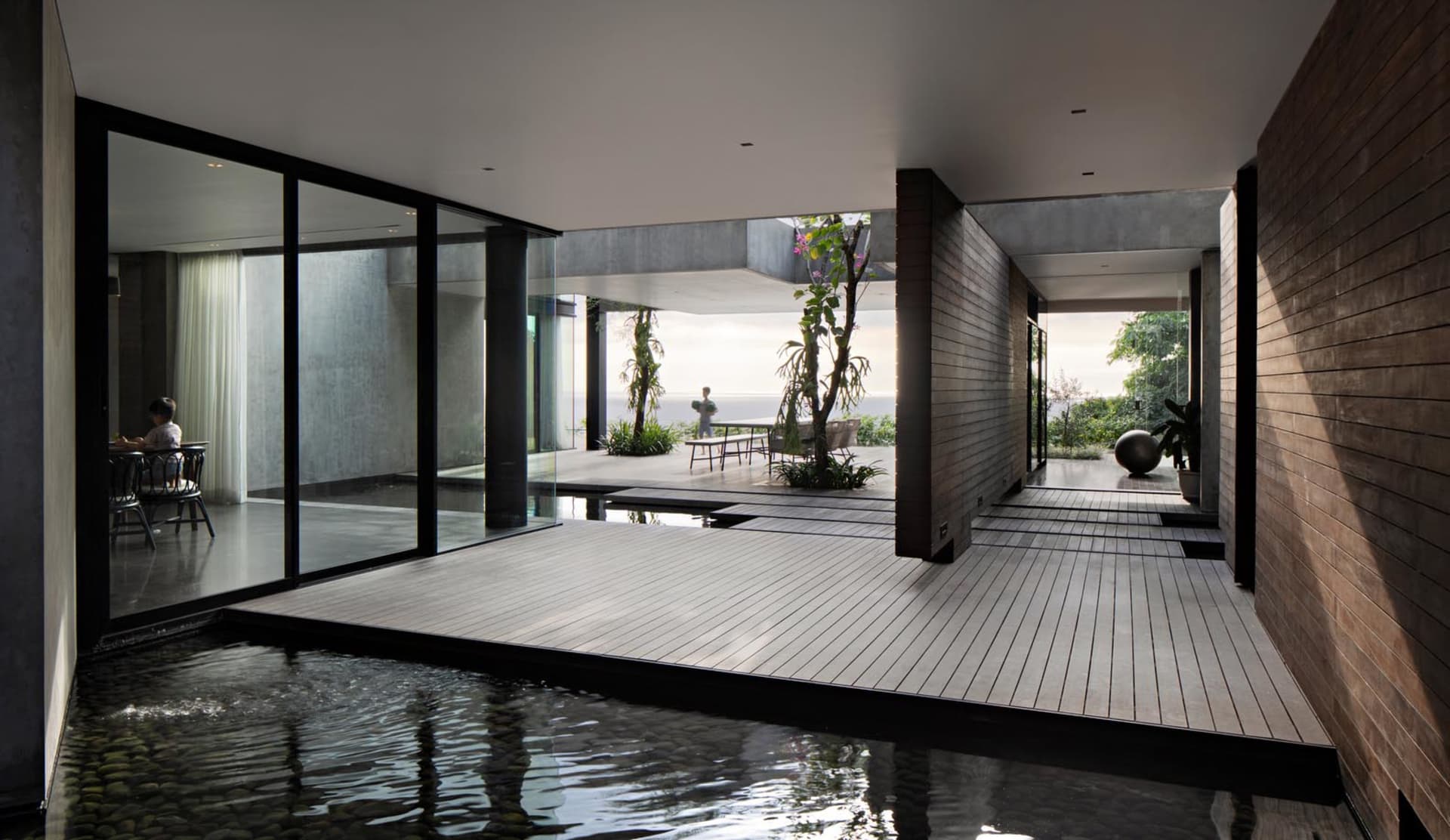 Modern residential living room with glass walls, wood floor, and concrete columns overlooking an outdoor space.