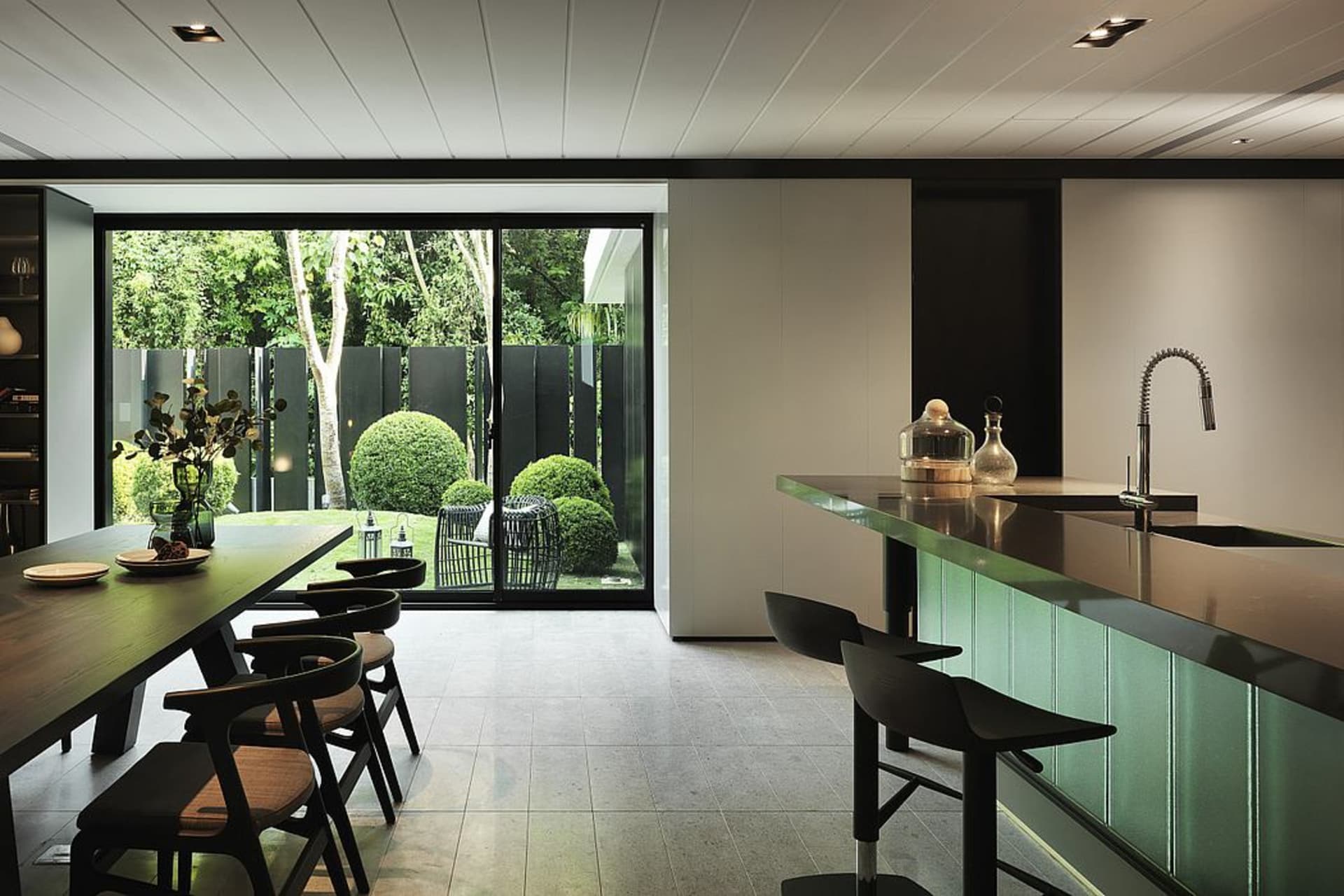 A modern kitchen with wooden flooring, a long wooden dining table, black chairs, and a kitchen island with a green countertop