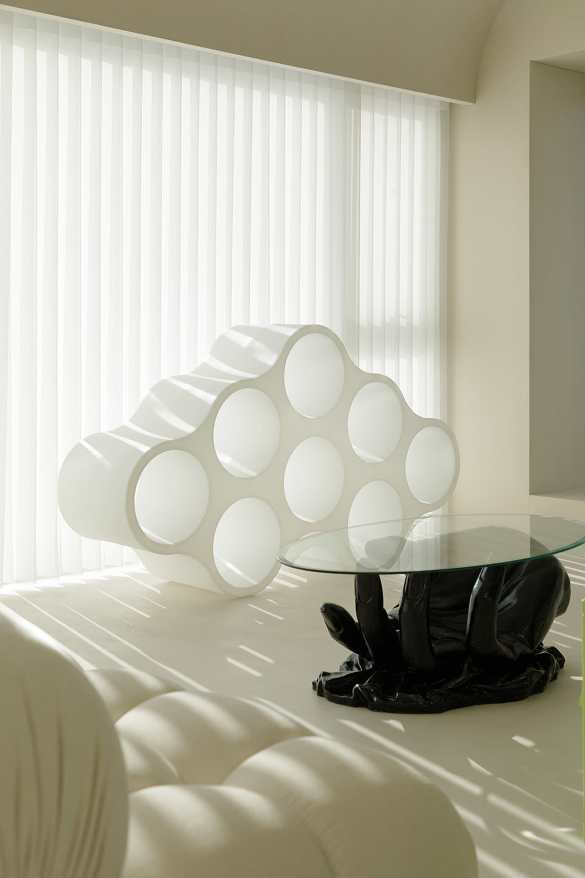 A minimalist space featuring a modern, glass coffee table with a hand-shaped base and a white, cloud-like structure with circ
