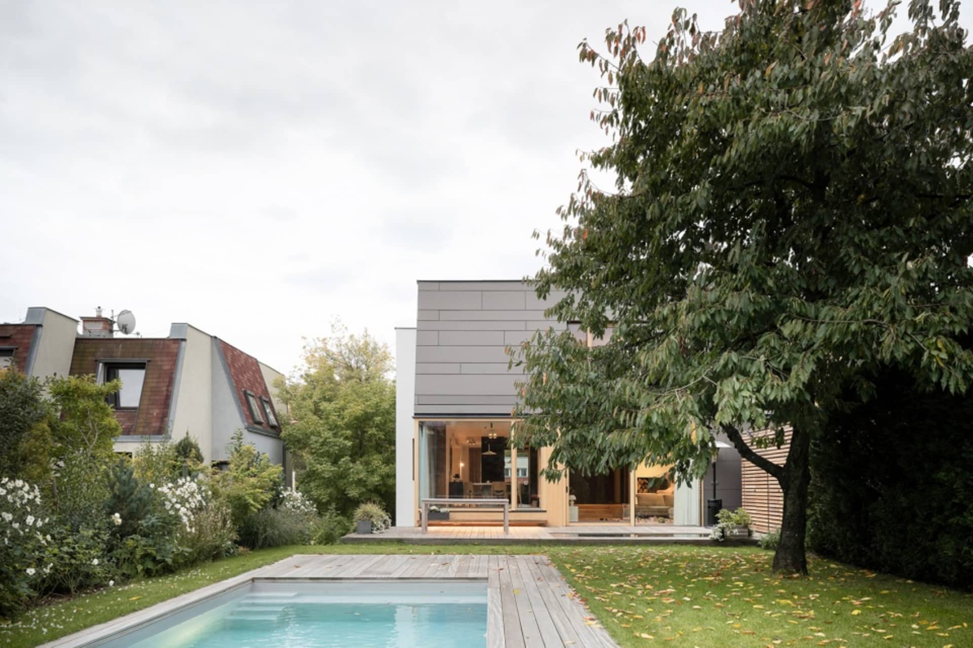 A modern residential backyard featuring a house with a pool and a large tree. The house has a minimalist design with wooden d