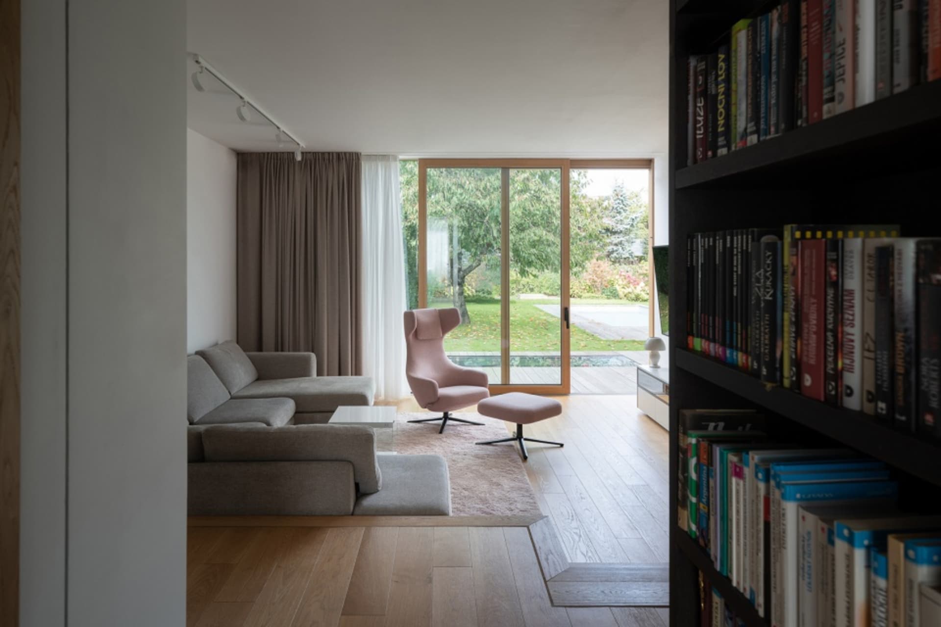 A modern living room featuring a bookshelf filled with books, a grey sectional sofa, and a pink lounge chair.