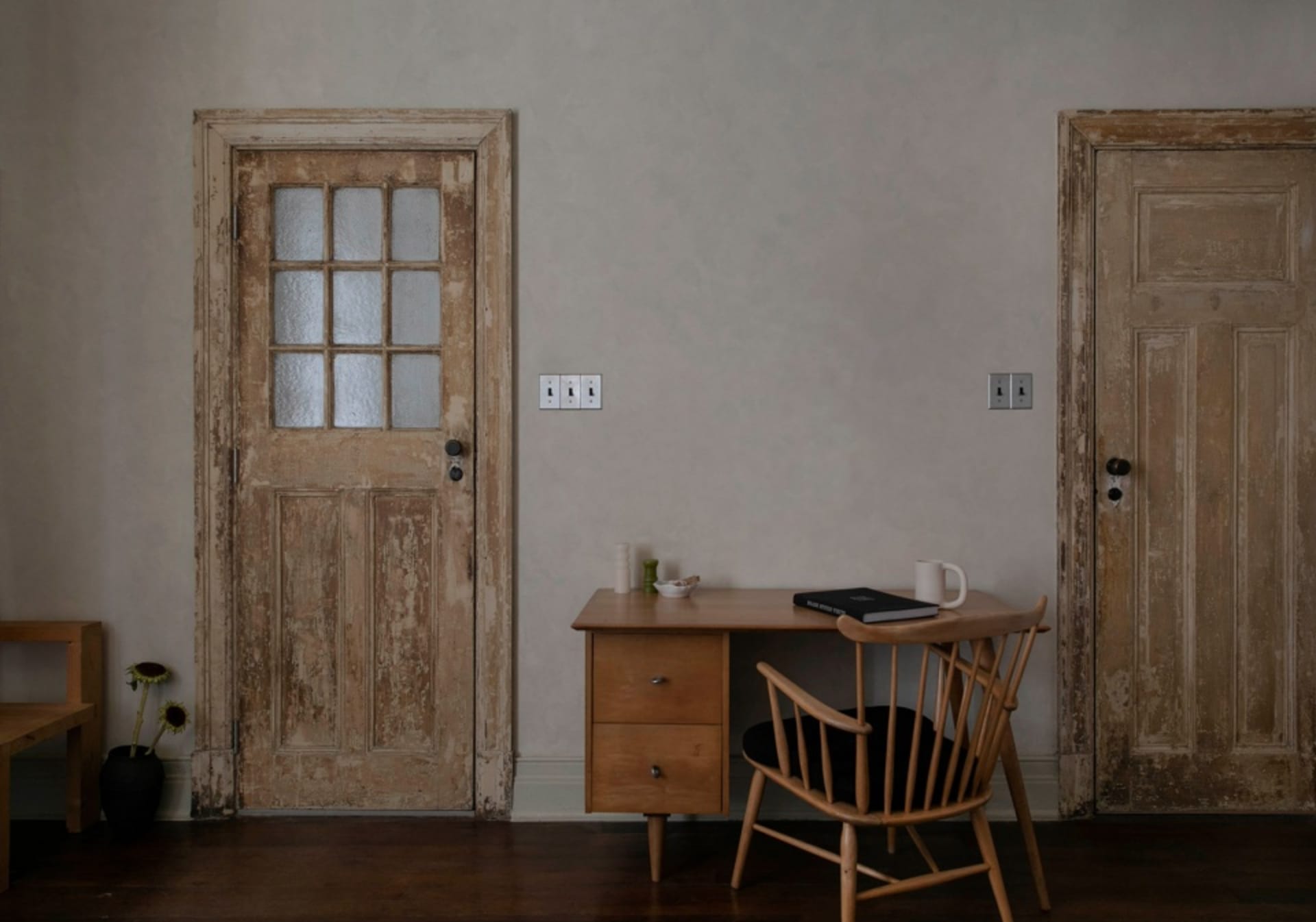 A minimalist interior featuring a wooden desk and chair against a plain wall, with two doors in the background.