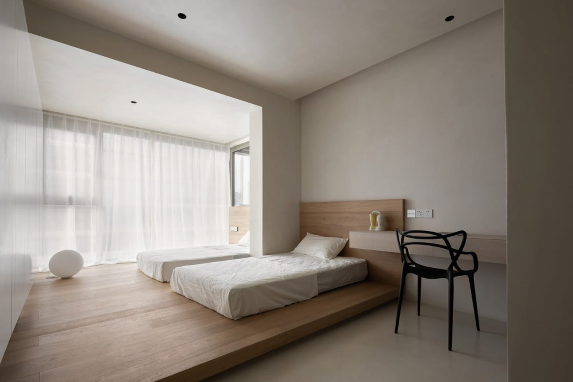 A minimalist bedroom featuring twin beds on a wooden platform with sheer curtains and a black chair against a white wall.