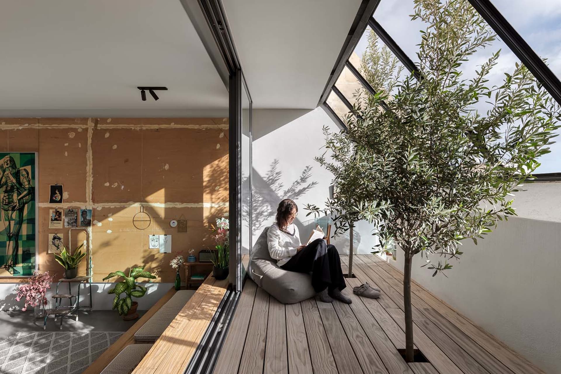 A woman sits on a cushioned chair reading a paper in a modern living space with wooden flooring and plants.